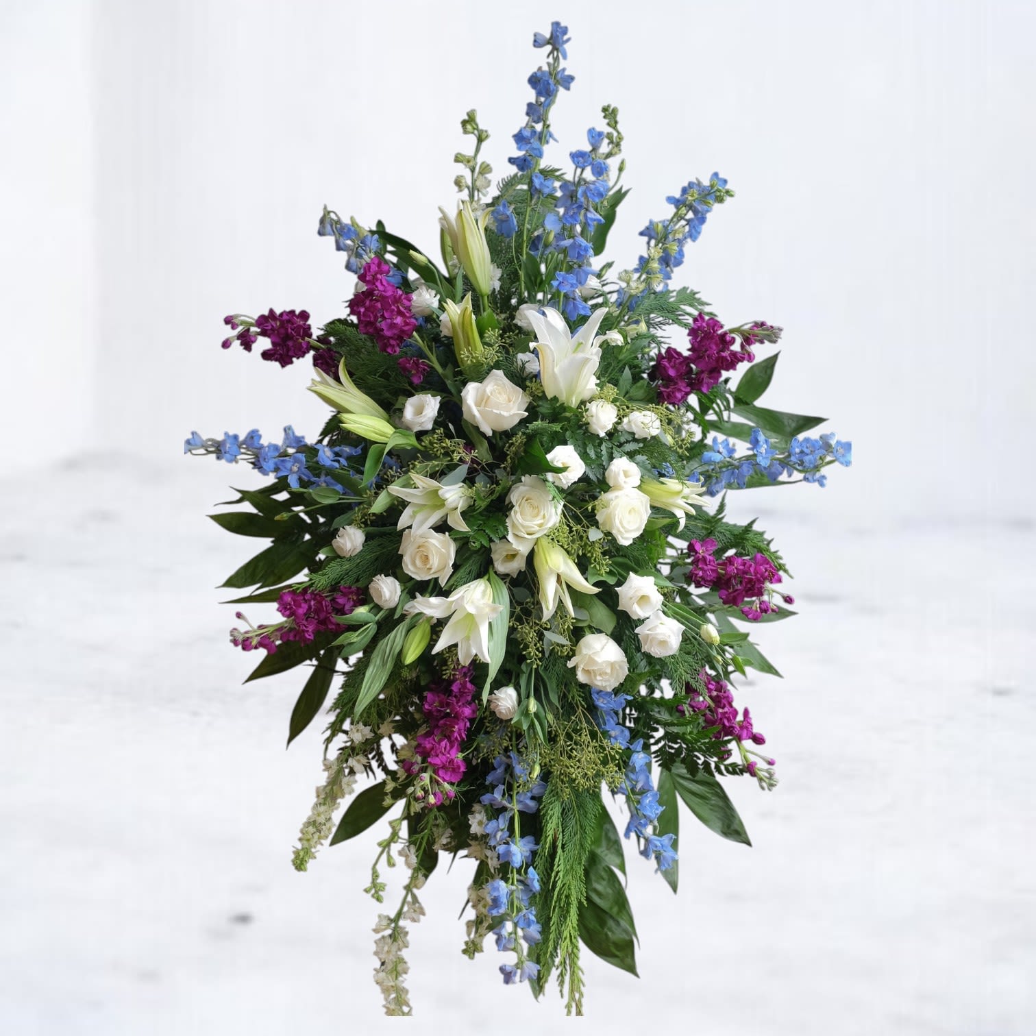 Vale Tribute Vale Tribute - Vale Tribute Standing Spray A breathtaking standing spray designed to honor a life with depth and dignity. The Vale Tribute features a serene blend of ivory roses, white oriental lilies, blue delphinium, and rich merlot stock, all arranged with lush greenery for striking contrast and natural movement. This elegant funeral flower arrangement is perfect for memorial services, church ceremonies, or graveside tributes. Available for same-day flower delivery to funeral homes across Houston, Magnolia, The Woodlands, and surrounding areas. Send a heartfelt message of peace, remembrance, and everlasting love with this custom sympathy spray. Easels are included with all standing sprays for effortless display at services.