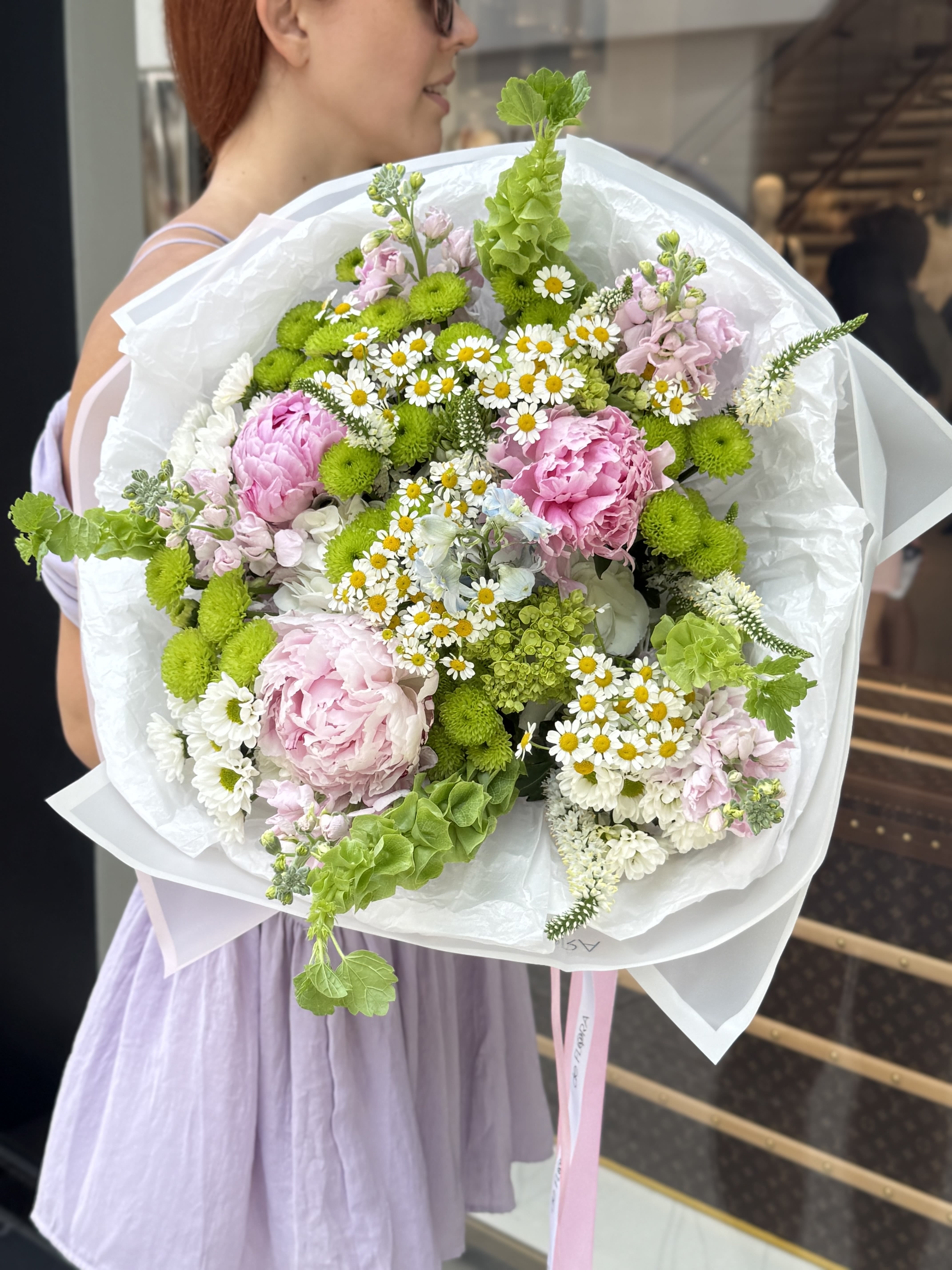 Spring Meadow - A fresh and whimsical bouquet that captures the essence of a blooming countryside. Spring Meadow features delicate pink peonies, fluffy lisianthus, soft matthiola, playful green button chrysanthemums, chamomile daisies, and bell-shaped bupleurum. The combination of gentle pink, crisp white, and vibrant green creates a harmonious and cheerful composition—perfect for birthdays, spring celebrations, or simply brightening someone’s day. Wrapped in elegant white paper and adorned with pastel ribbons from deFLORA, this bouquet is a breath of fresh air in floral form.