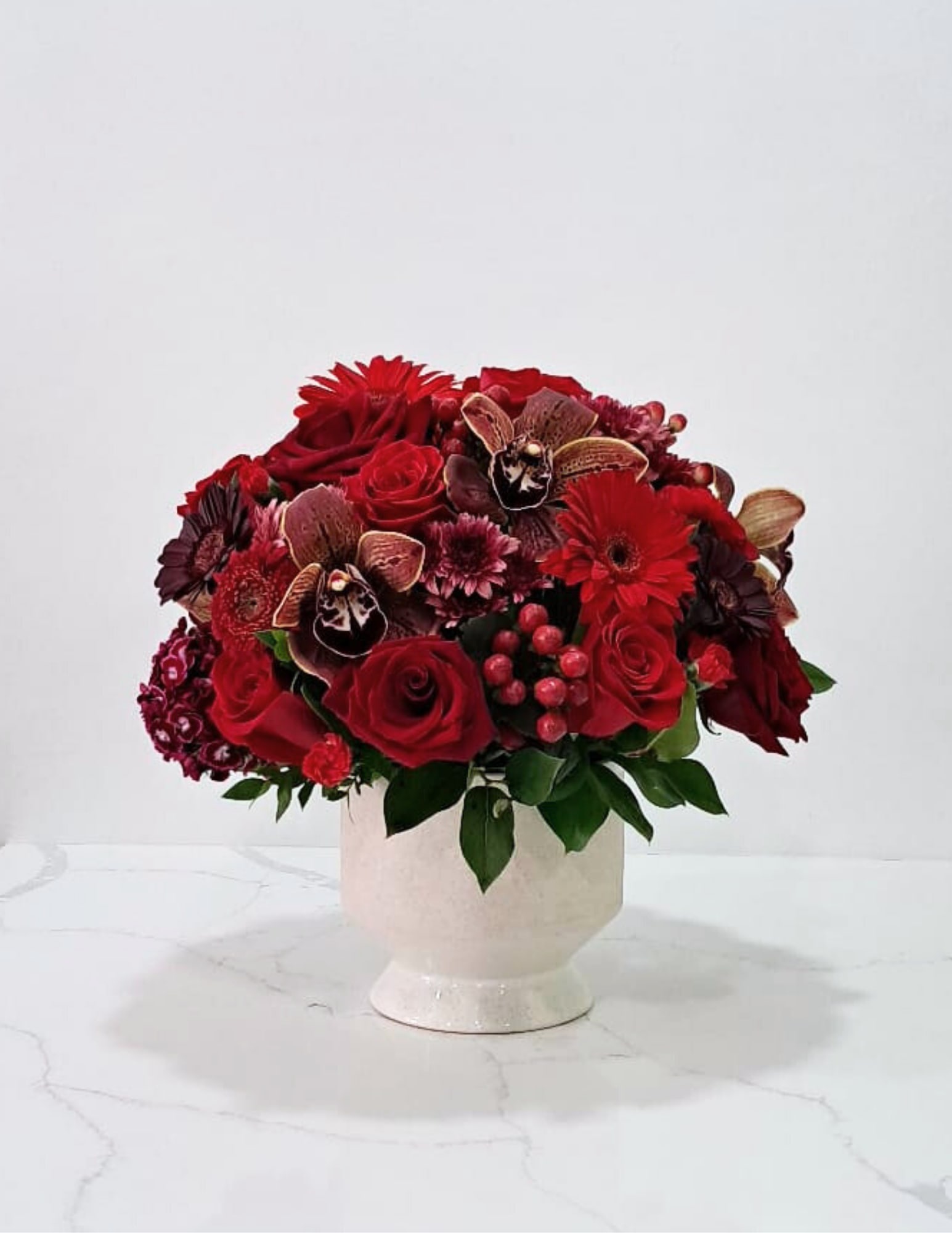 Black Cherry Black Cherry - This gorgeous arrangement has mini gerbera daisies, red roses, mums, hypericums, cymbidium orchids, gerbera daisies, mini carnations and sweet williams in a ceramic vase.