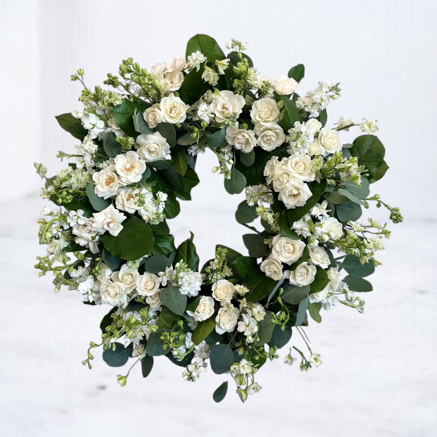 Whispervale Tribute Funeral Wreath - A graceful all-white wreath that speaks volumes in silence. The Whispervale Tribute Funeral Wreath features delicate white roses, ruffled stock, and soft sprays of lisianthus, artfully arranged among rich greenery for a refined and timeless look on a 15&quot; wreath. Designed as a symbol of peace and remembrance, this wreath offers comfort and reverence in moments of loss.  Ideal for funeral services or memorials in Houston, Magnolia, Tomball, and surrounding areas. Same-day delivery available to local funeral homes.