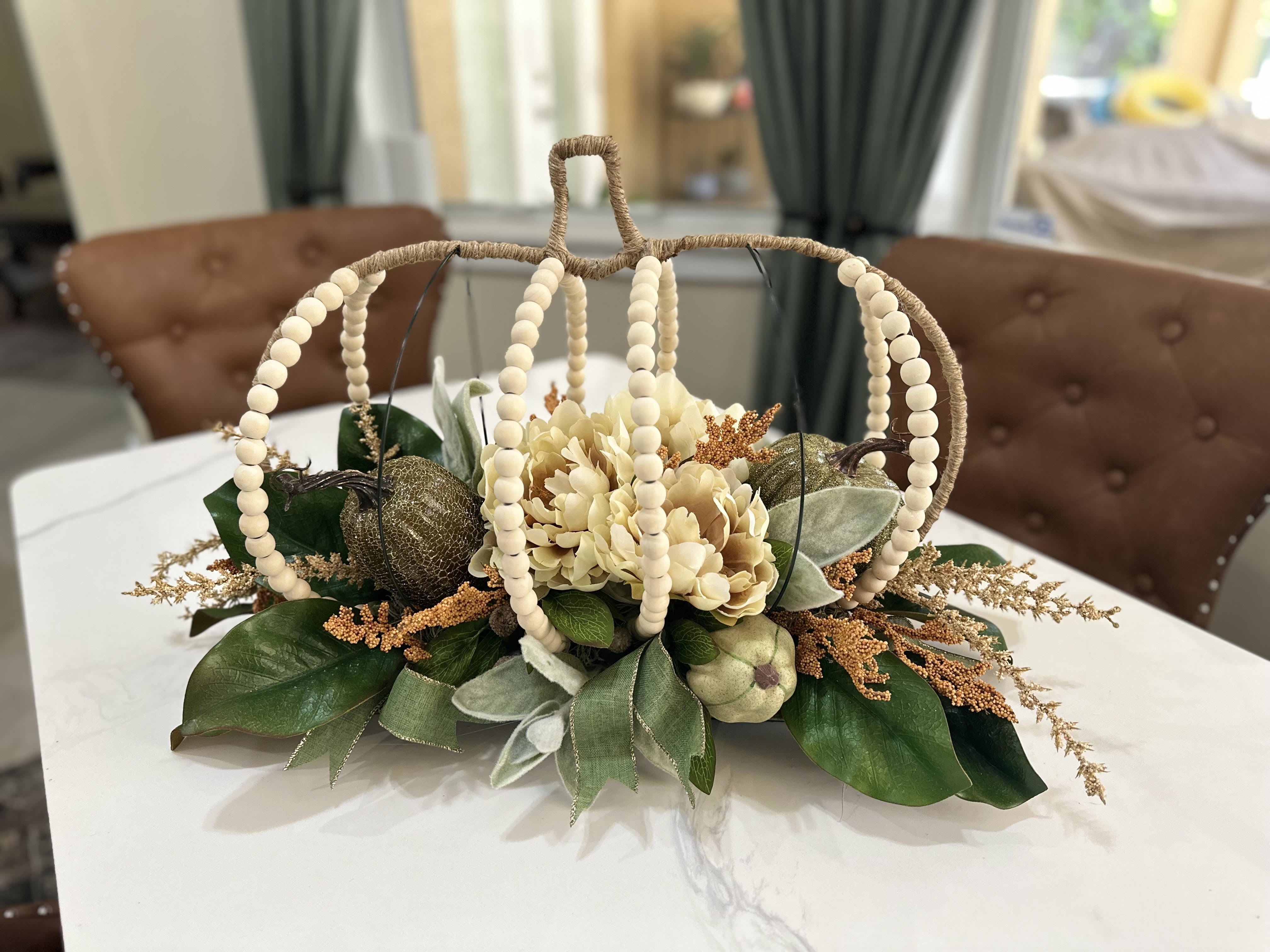 Harvest Elegance Centerpiece -  Bring the warmth of autumn into your home with this stunning Autumn Harvest Centerpiece. Crafted with cream-colored flowers, lush green leaves, and decorative gold pumpkins, this arrangement is nestled in a charming watted basket adorned with wooden beads. Perfect for dining tables, entryways, or any space wanting a touch of seasonal elegance. Adds a cozy, rustic charm to your fall decor.