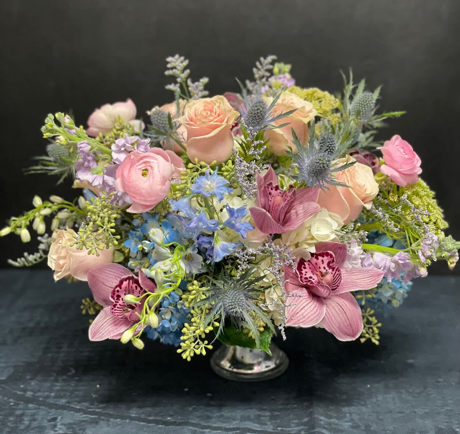 A Whisper of Pastels - A soft, romantic palette that evokes elegance and tranquility. This arrangement features Blue Hydrangeas, Lavender Stock, Light Pink Roses, Light Pink Ranunculus, Pink Cymbidiums, Blue Delphinium, Blue Thistles, Caspia, and Eucalyptus — all artfully arranged in a classic Silver Compote vase.
