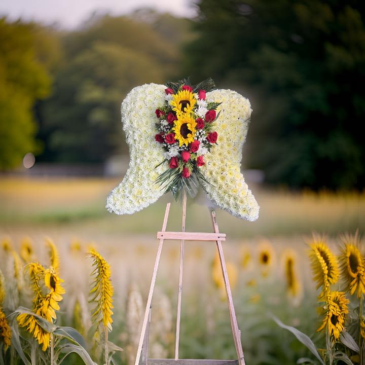 Angel Wings Sympathy Standing - A heartfelt floral tribute in the shape of angel wings, symbolizing peace, love, and eternal rest. This elegant arrangement is crafted from a bed of soft white flowers, beautifully accented with vibrant sunflowers, red roses, and touches of greenery in the center. Displayed on a wooden easel, it’s a graceful way to express condolences and honor a life well lived.  Perfect for memorials, funerals, or celebration of life services.