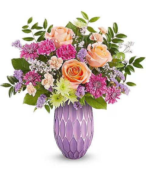 Teleflora's Pretty Glow Bouquet - Make her Mother's Day. Shine with the Pretty Glow Bouquet, featuring a stunning mix of roses, carnations, and chrysanthemums, beautifully presented in a sculpted glass vase with a purple mercury-inspired finish. A perfect blend of elegance and charm to brighten her special day.  Peach roses, fuchsia carnations, peach miniature carnations, green cushion spray chrysanthemums, purple cushion spray chrysanthemums, purple sinuata statice, and pink limonium are artfully arranged with huckleberry and salal in the Pretty Glow vase, creating a stunning Mother's Day bouquet.