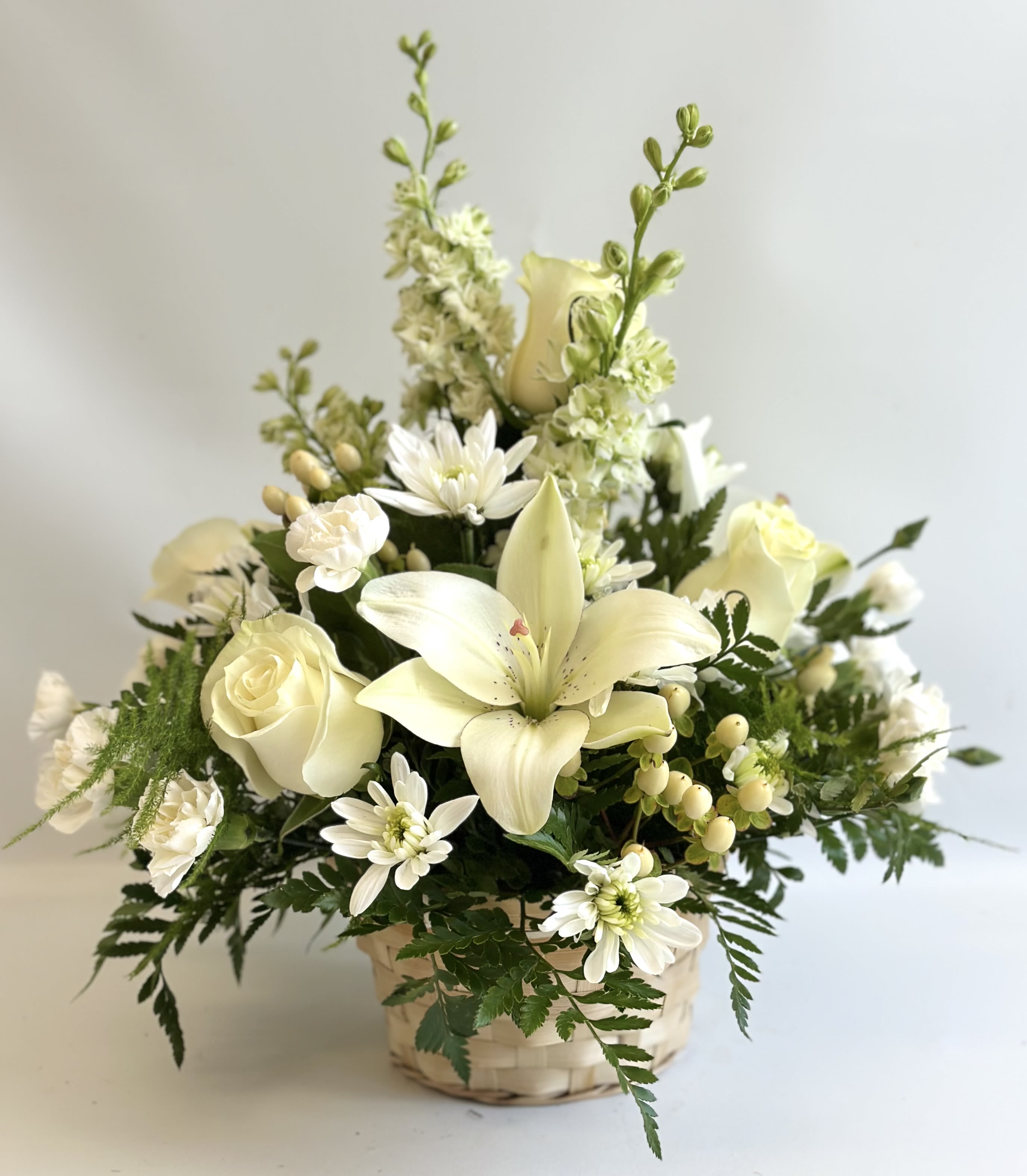 Winter’s Embrace by Barb’s Flowers - An all white arrangement of Asiatic Lilies(non fragrant), Roses, Cushion Mums, Mini Carnations, Hypericum Berries and Larkspur or Snapdragons.  This is an all around design. In a basket. 