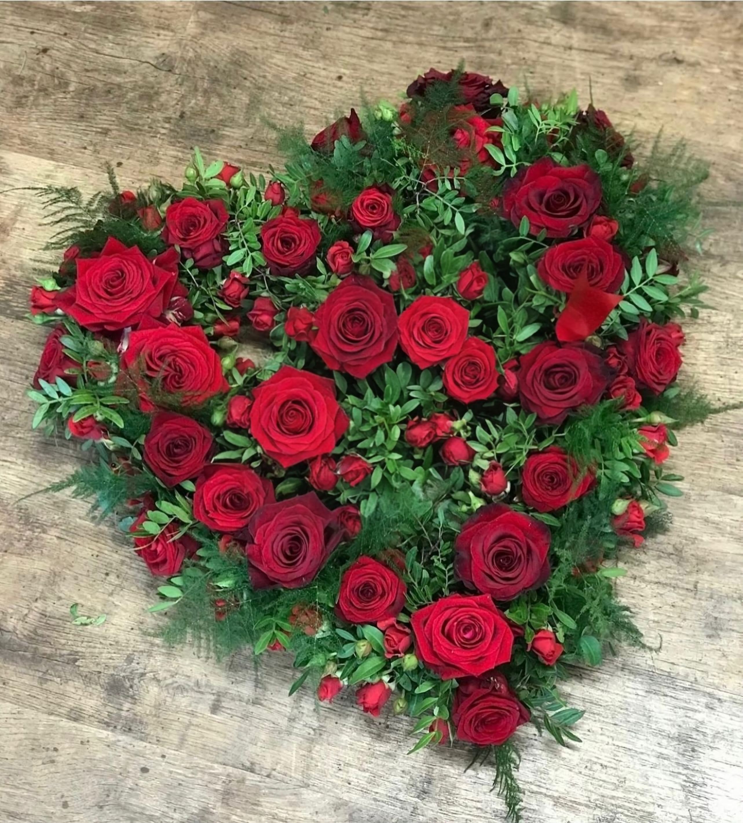 Heartfelt Devotion - This captivating heart-shaped arrangement is a timeless expression of love and devotion. Crafted with rich, velvety red roses, symbolizing deep passion and romance, it also features delicate spray roses for added texture and depth. The lush green fern leaves and ruscus gracefully surround the blooms, accentuating the heart's shape while providing a natural contrast. Perfect for conveying heartfelt emotions, this arrangement is ideal for romantic gestures, weddings, or memorial tributes, creating a powerful statement of enduring love and admiration.  Red Roses: The most prominent flowers in the arrangement are the red roses, symbolizing love, passion, and romance.    Spray Roses: In addition to the larger red roses, there are smaller red spray roses scattered throughout. Spray roses are smaller blooms that grow in clusters on a single stem, and they complement the larger roses while adding texture to the arrangement.  Fern Leaves: The arrangement includes fern leaves, which have a feathery, delicate appearance.   Ruscus or Myrtle: There is greenery with elongated, rounded leaves, likely either ruscus or myrtle.        