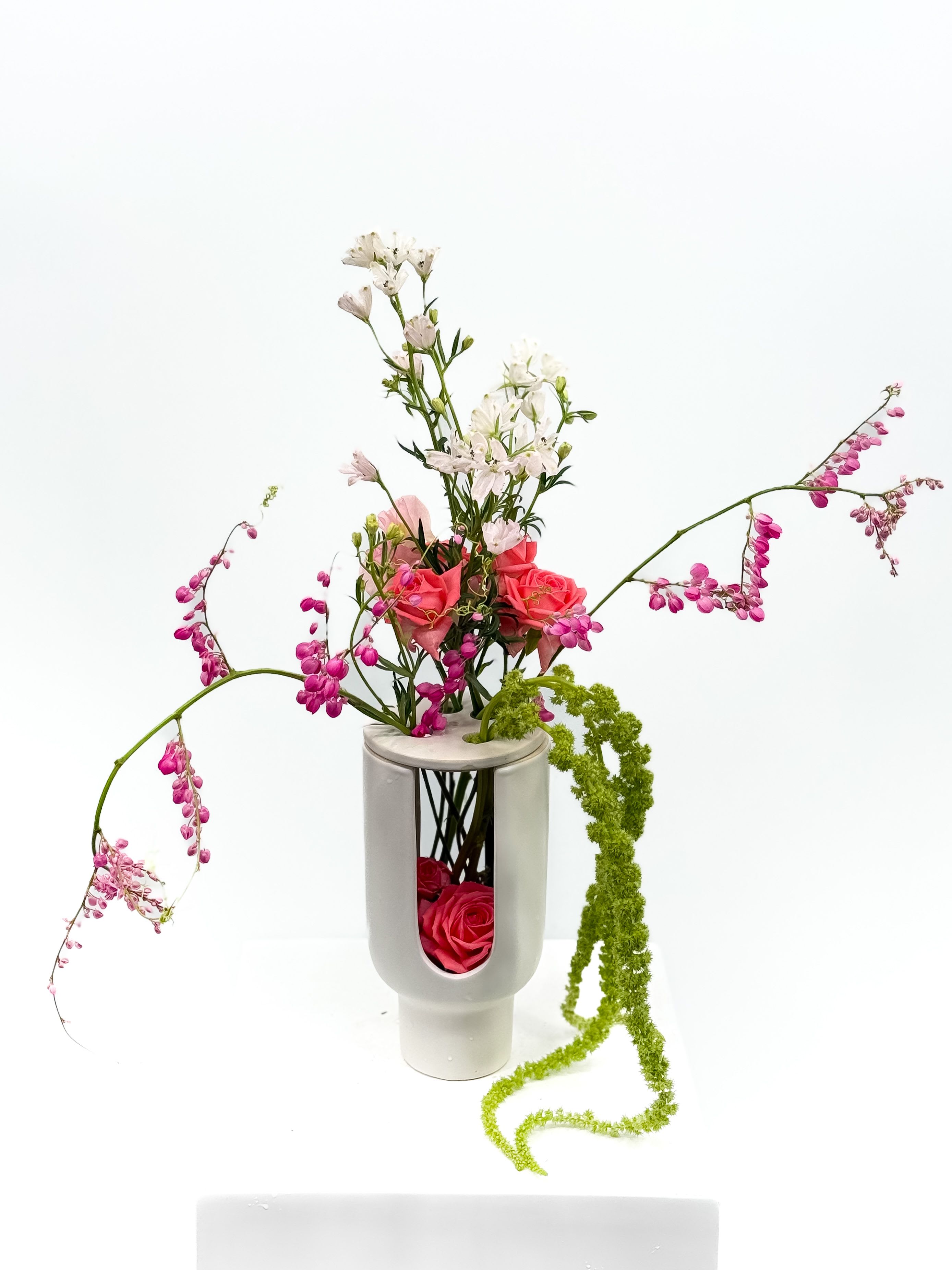 Zara Zara - This striking arrangement blends sculptural design with garden whimsy. Featuring cascading pink blooms, delicate whites, and vibrant greens, it’s all brought to life by a ceramic vase with a bold cut-out—showcasing a hidden cluster of roses within. A true conversation piece that turns florals into art.