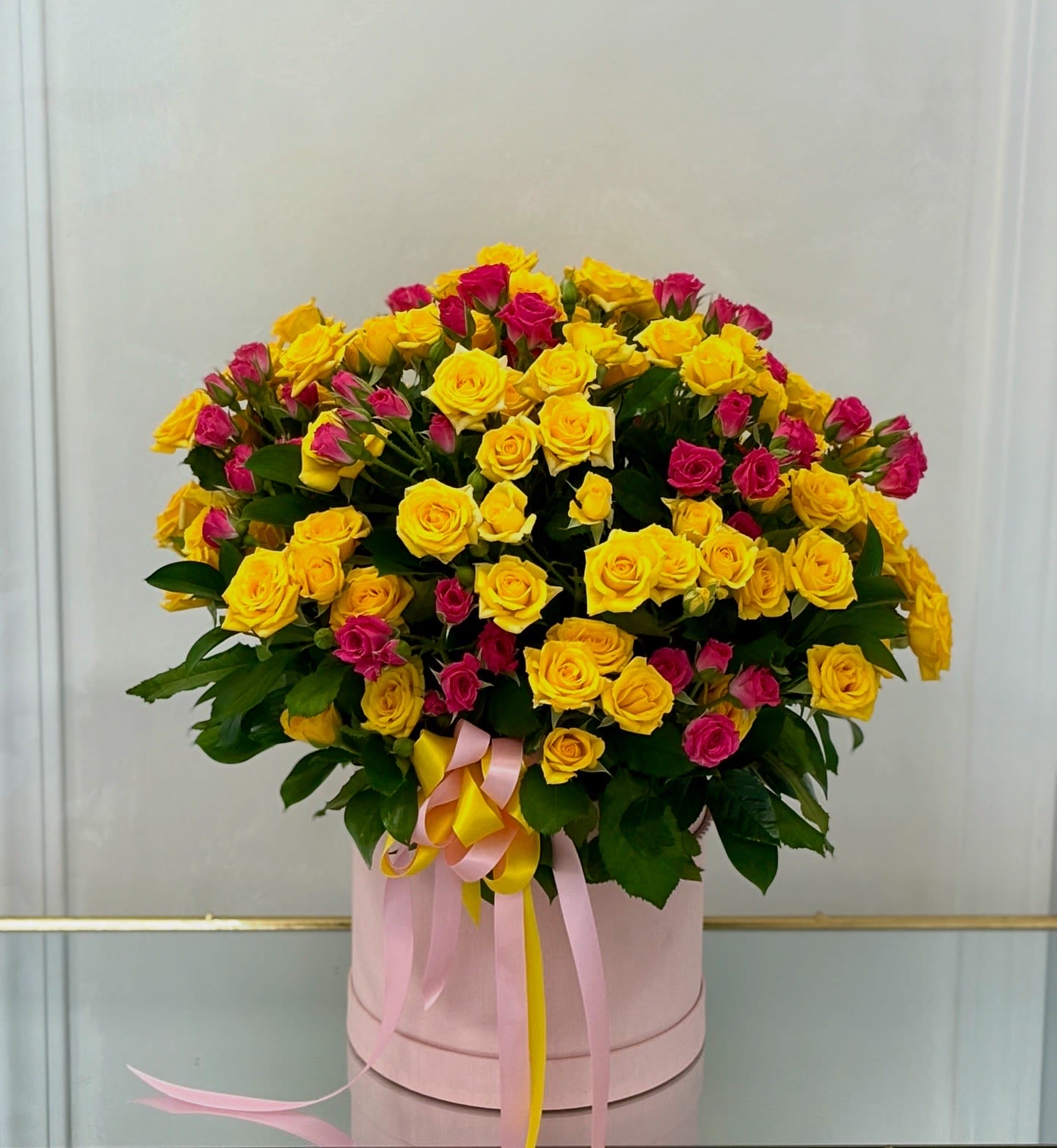 Sunset Kisses By VLM Flowers - Overflowing with joy, Sunset Kisses features an abundant mix of golden yellow and hot pink spray roses arranged in a stylish round hatbox. Accented with playful pink and yellow satin ribbons, this cheerful design radiates warmth, happiness, and celebration—perfect for birthdays, congratulations, or brightening someone’s day just because.