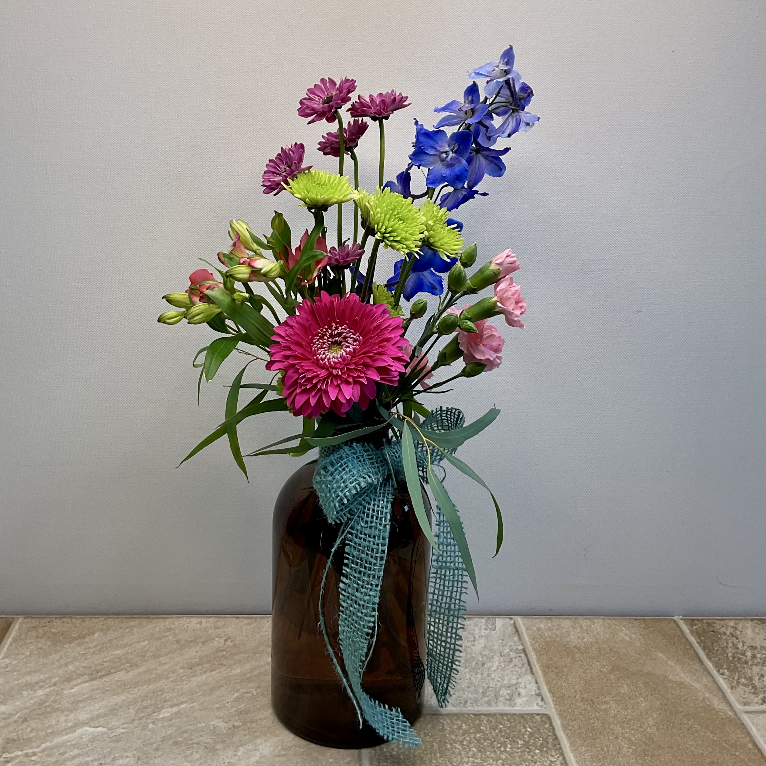 Wildflower Whimsy Wildflower Whimsy - This playful and eclectic floral arrangement brings a touch of countryside charm with a burst of vibrant color. Set in a rustic amber glass jar and tied with a teal burlap ribbon, the bouquet features a lively mix of blooms. A bright pink gerbera daisy takes center stage, complemented by lime green athos and lavender micro button mums, soft pink mini carnations, pink alstroemeria, and striking blue delphinium. The asymmetrical design and varying heights create a casual, garden-picked look that's both cheerful and modern. Ideal for brightening a kitchen nook, office space, or sending a little joy someone's way. Approximate size: 8" wide x 20" high Primary flowers: daisy, mum, carnation, lily, delphinium