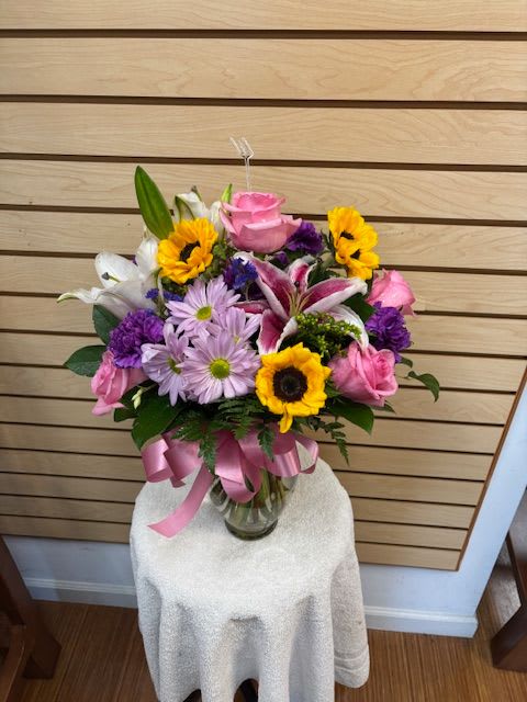 Chances - Mixed cut arrangement with stargazer lilies, white oriental lily, pink roses, sunflowers, purple carnation, lavender daisy, and purple statice in a vase with a bow