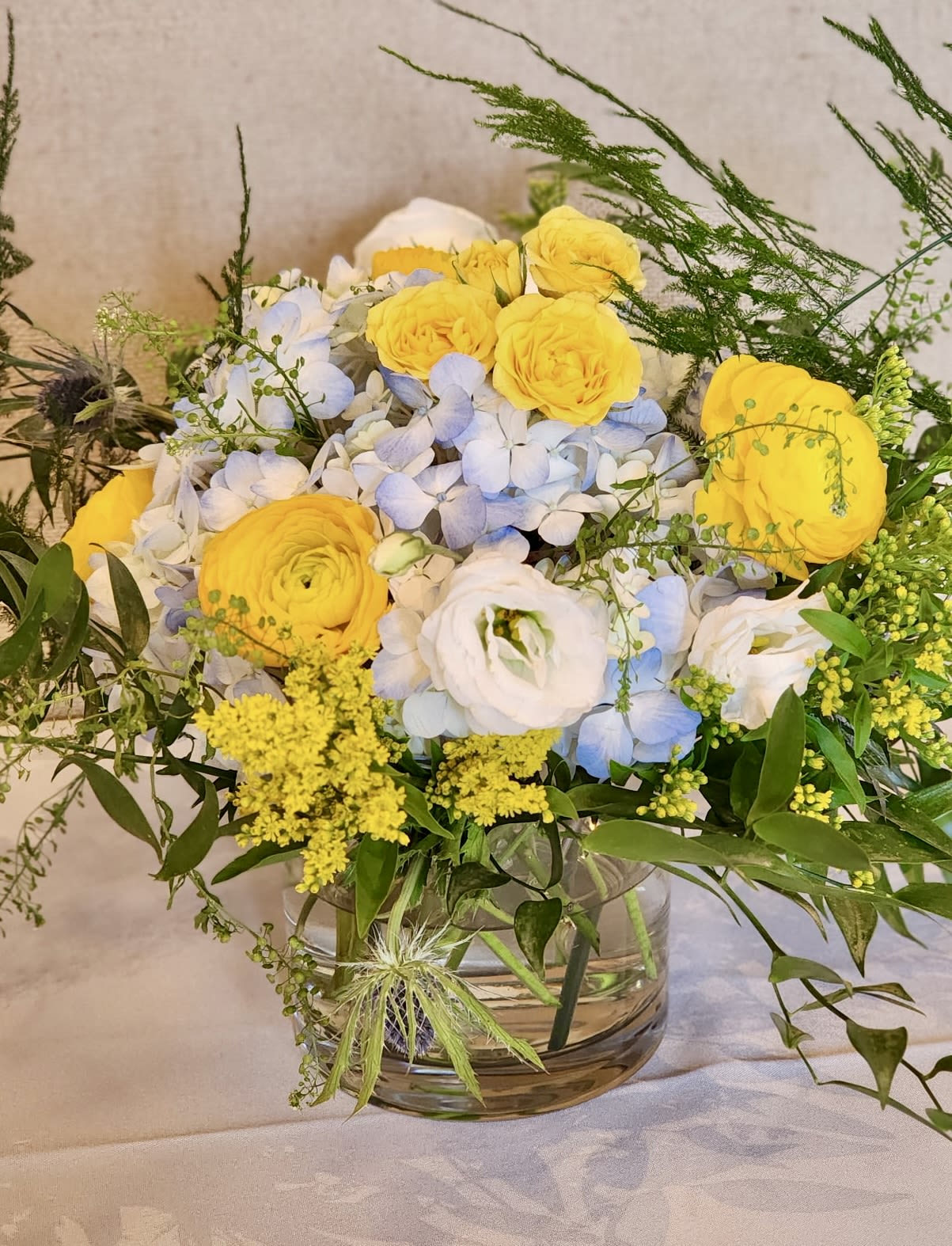 Belle Belle - This cool and serene centerpiece is filled with blue, white and yellow blooms. It’s a beautiful and timeless. Glass vase included. Approximately 12x12 in size Every effort will be made to recreate your chosen design. Please note that from time to time due to product availability and quality we may need to make substitutions. We will make sure to keep the original design in mind when making substitutions.