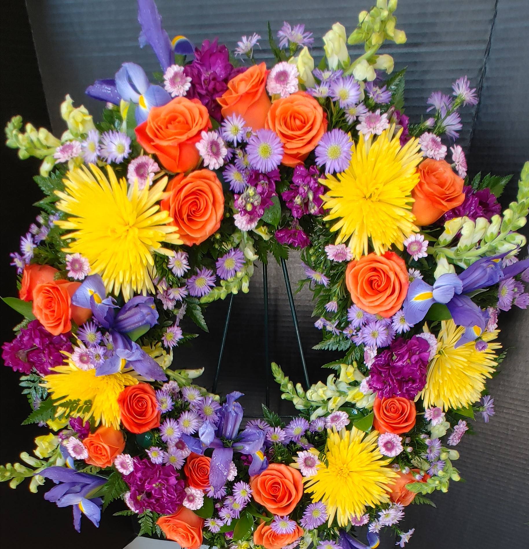 A Colorful Life Wreath - A bright colorful mixture to include, Orange Roses, Yellow Snaps, Lavender buttons, Purple Monte, Purple Iris, Fuchsia Stock, Yellow Fuji Mums and mixed greenery