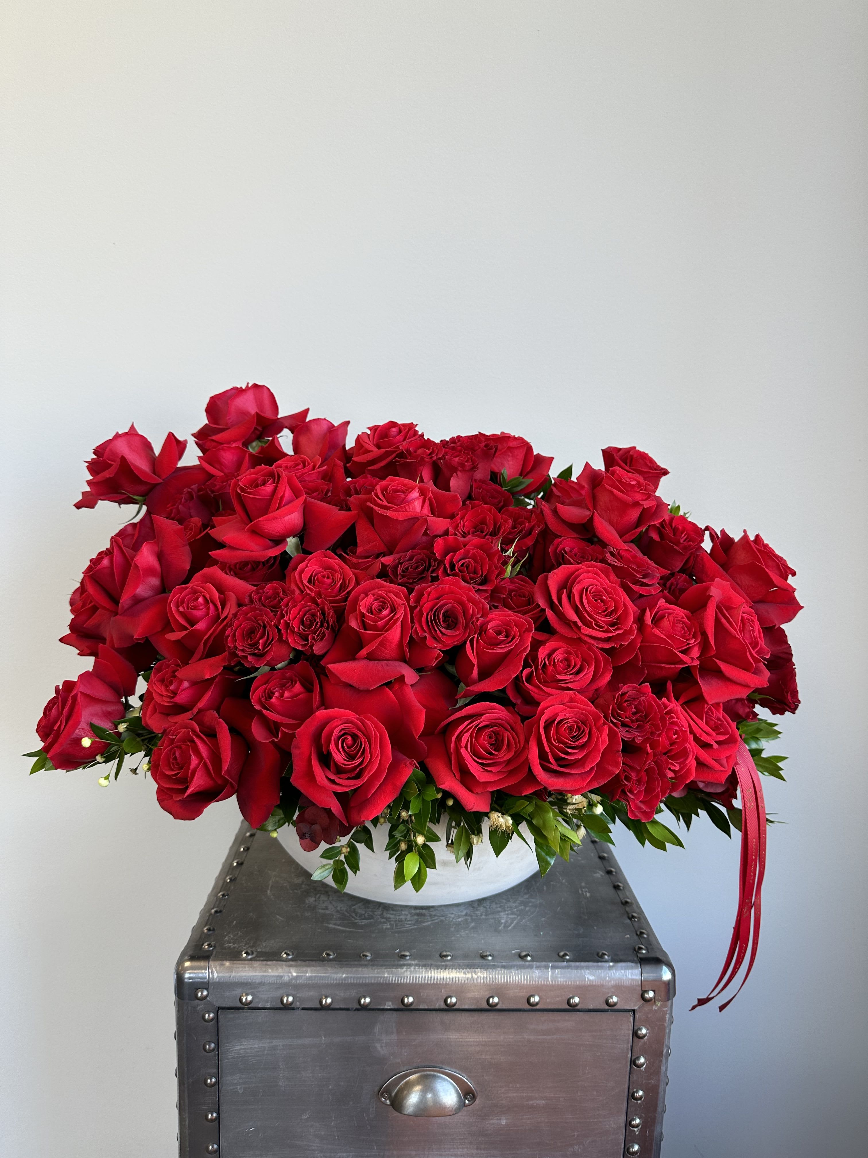 Brooklyn - A stunning arrangement of lush red roses elegantly presented in a low white ceramic bowl, accented with rich greenery and finished with a red ribbon for a bold, romantic statement.