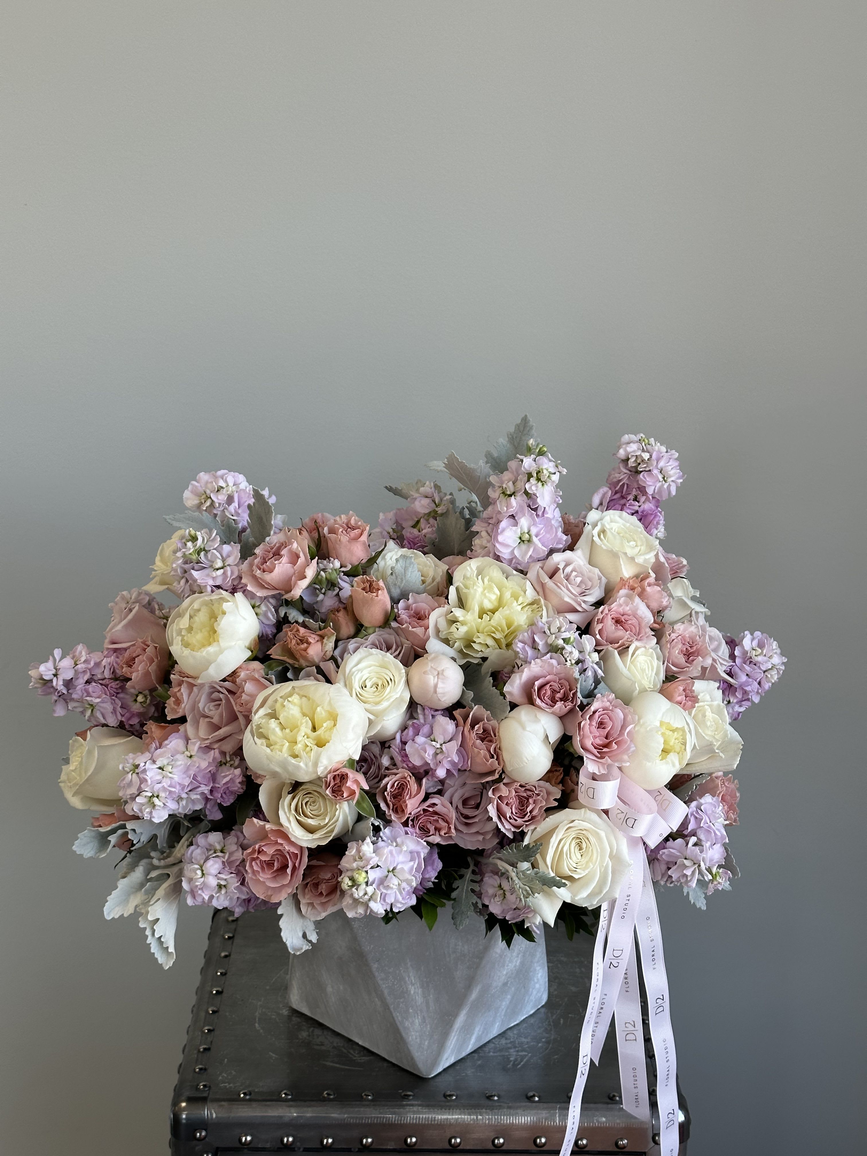 Catalina - A soft and romantic arrangement of pastel blooms featuring ivory peonies, blush roses, and lilac stock, artfully arranged in a geometric stone-grey vase and finished with elegant ribbon detailing—perfect for celebrating love, grace, and timeless beauty.
