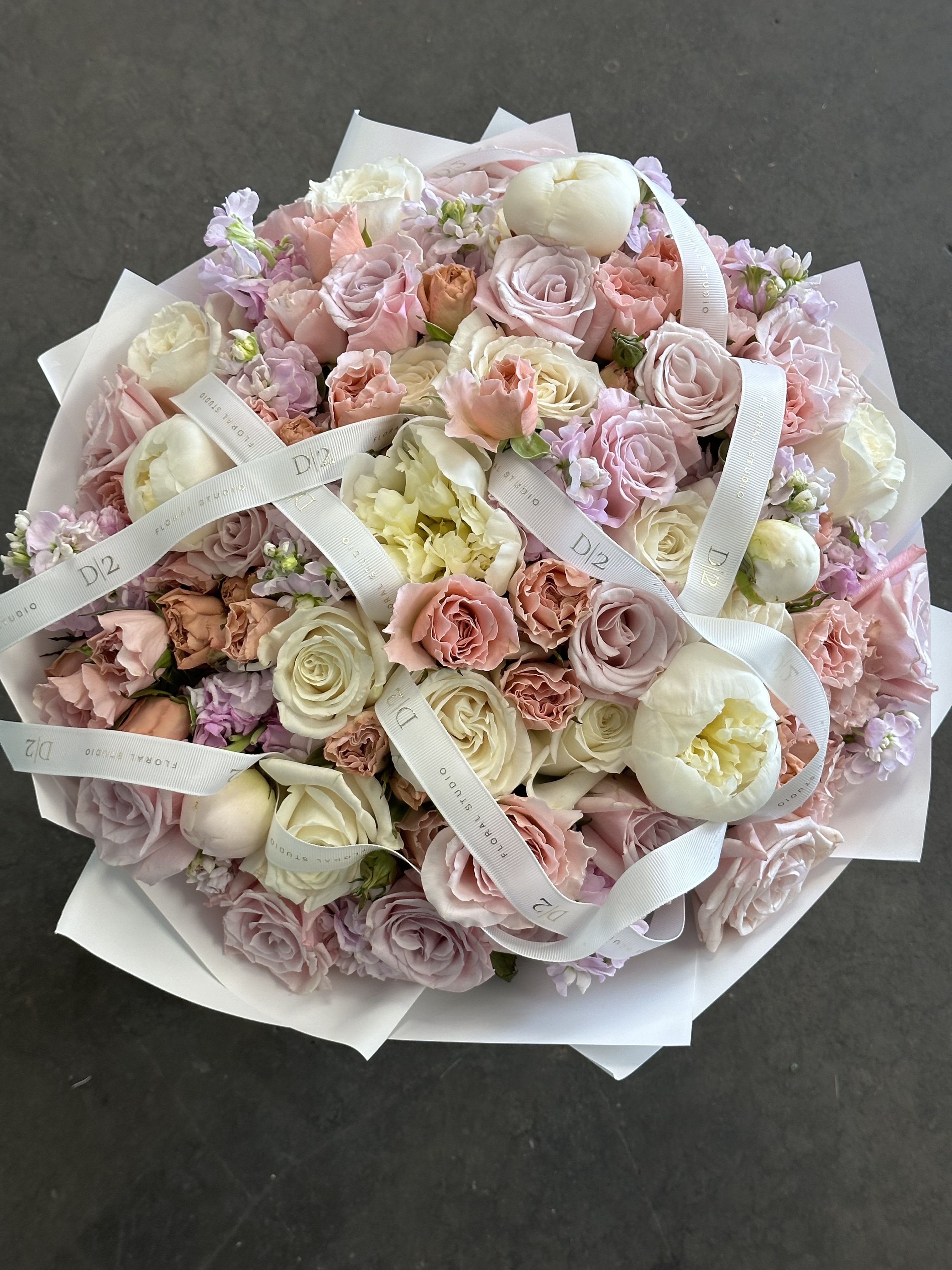 Alina Alina - A charming round bouquet overflowing with soft blush, ivory, and peach blooms, including roses, peonies, and delicate stock, wrapped in crisp white paper and tied with elegant branded ribbon—ideal for romantic gestures or refined celebrations.