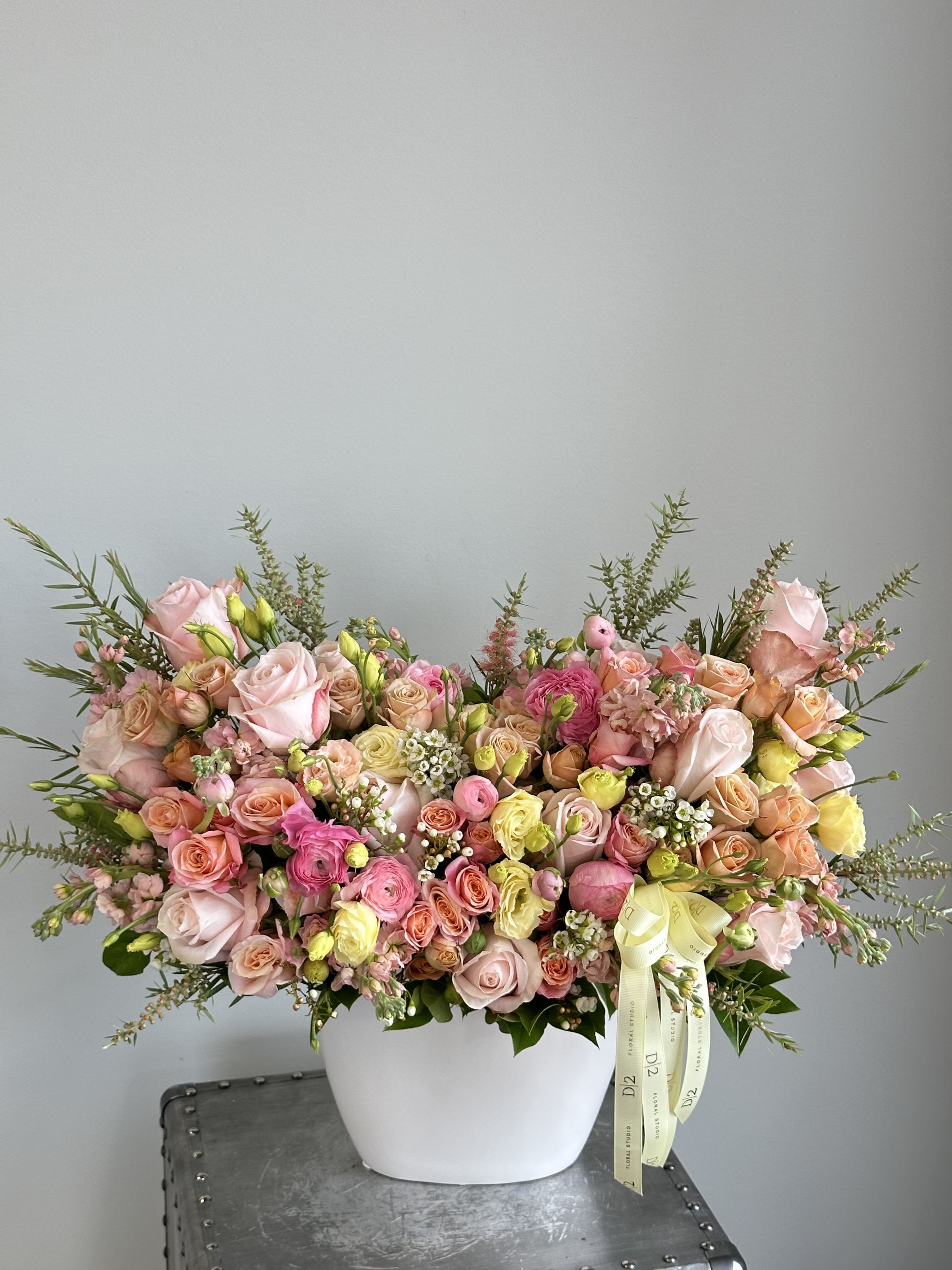 Summer Monica - A joyful and colorful floral arrangement bursting with pastel roses, ranunculus, spray roses, and wildflower accents in shades of pink, peach, yellow, and cream. Arranged in a sleek white vase and finished with a soft yellow ribbon—perfect for birthdays, spring celebrations, or cheerful gifts.