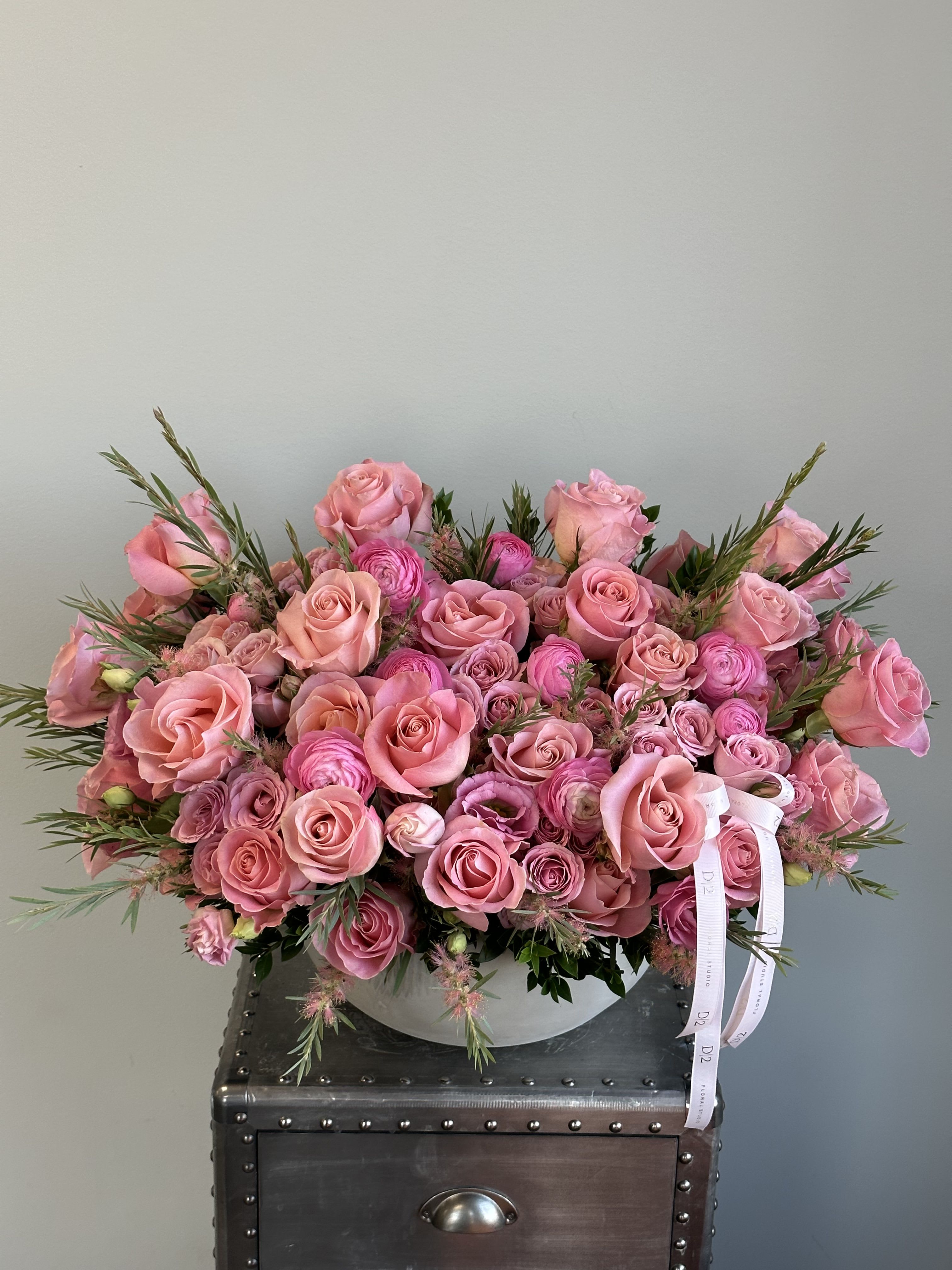 Daphne Daphne - radiant arrangement of vibrant pink roses and ranunculus in varying shades, accented with soft greenery and styled in a sleek white ceramic bowl. Finished with signature D|2 ribbons, this bouquet is a bold and beautiful expression of love and joy.