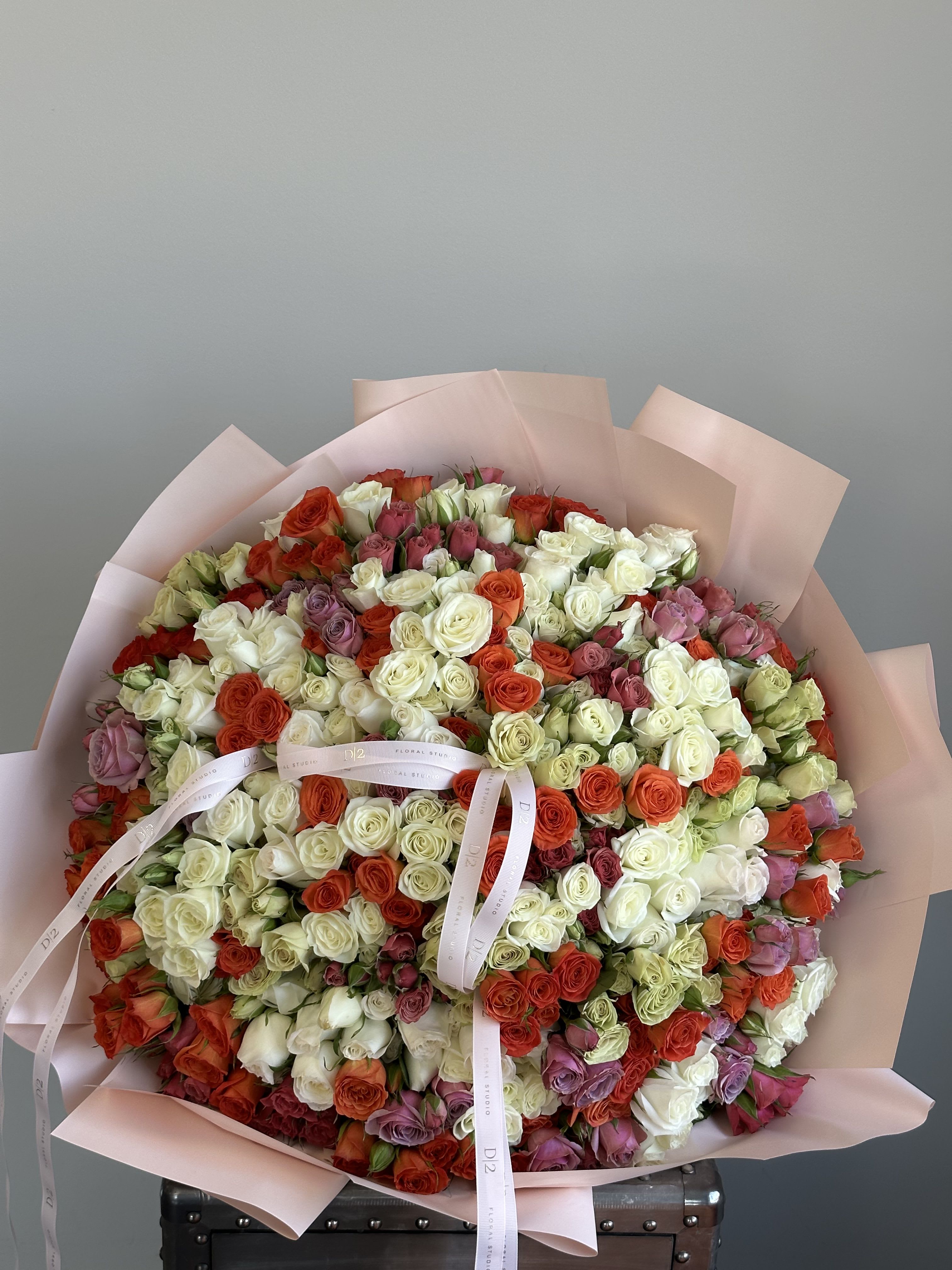 Adaline Adaline - An extravagant round bouquet bursting with vibrant mini roses in shades of white, red, orange, pink, and lavender. Artfully wrapped in blush pink paper and finished with signature D|2 ribbons—perfect for making a grand, colorful statement on any special occasion.