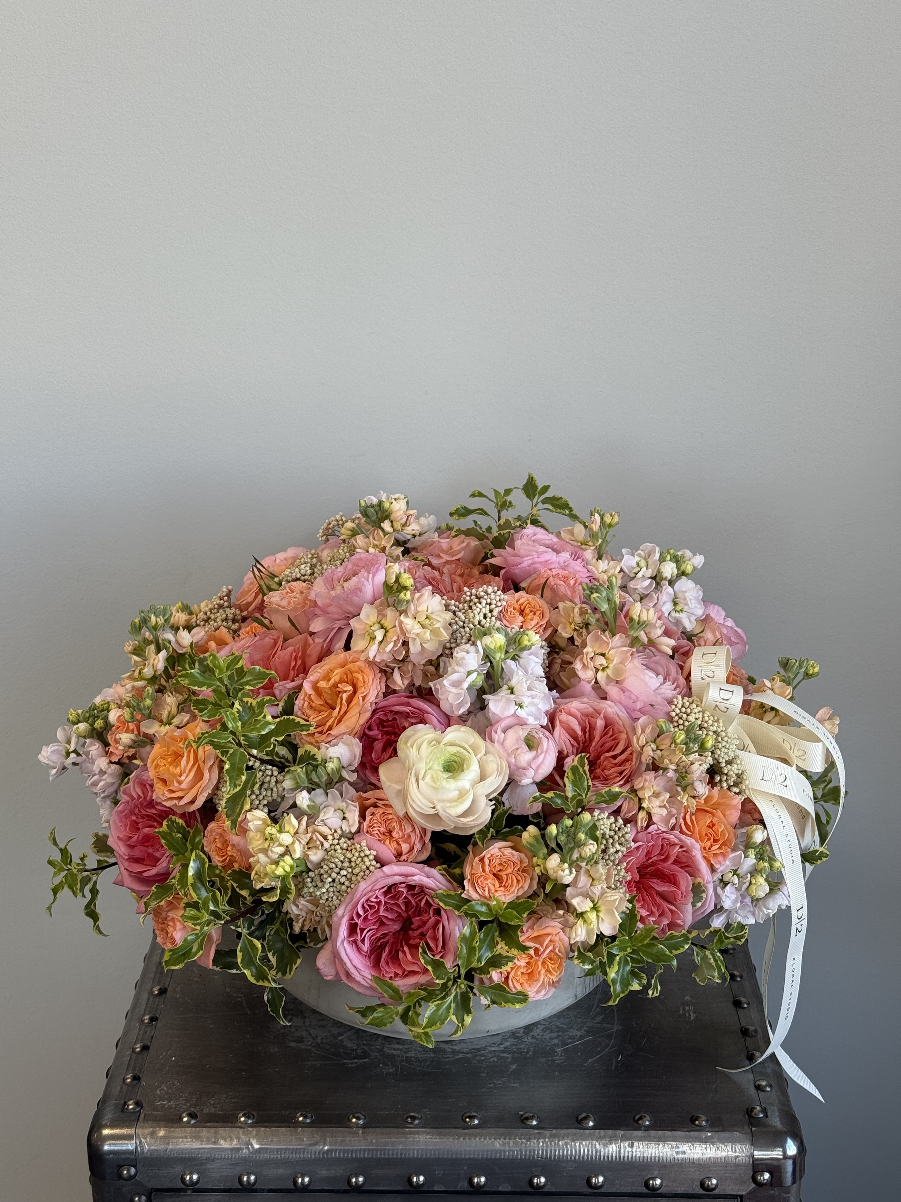 Nyla - A lush and vibrant floral arrangement featuring garden roses, ranunculus, and stock in shades of peach, coral, blush, and ivory, accented with textured fillers and trailing greenery. Presented in a round stone vase and tied with elegant D/2 ribbons—perfect for brightening any space or occasion.