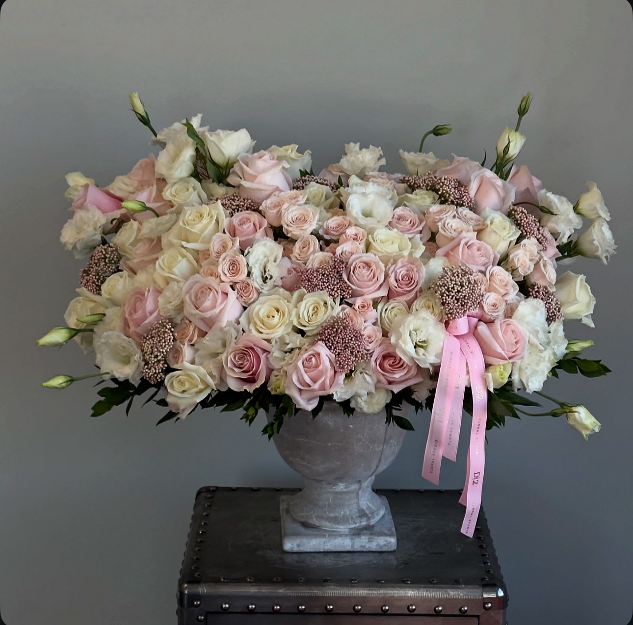 Kaia Kaia - An opulent and romantic arrangement of blush and ivory roses, lisianthus, and textured pink fillers, elegantly displayed in a classic stone urn and finished with a soft pink ribbon—perfect for weddings, anniversaries, or graceful sympathy tributes.