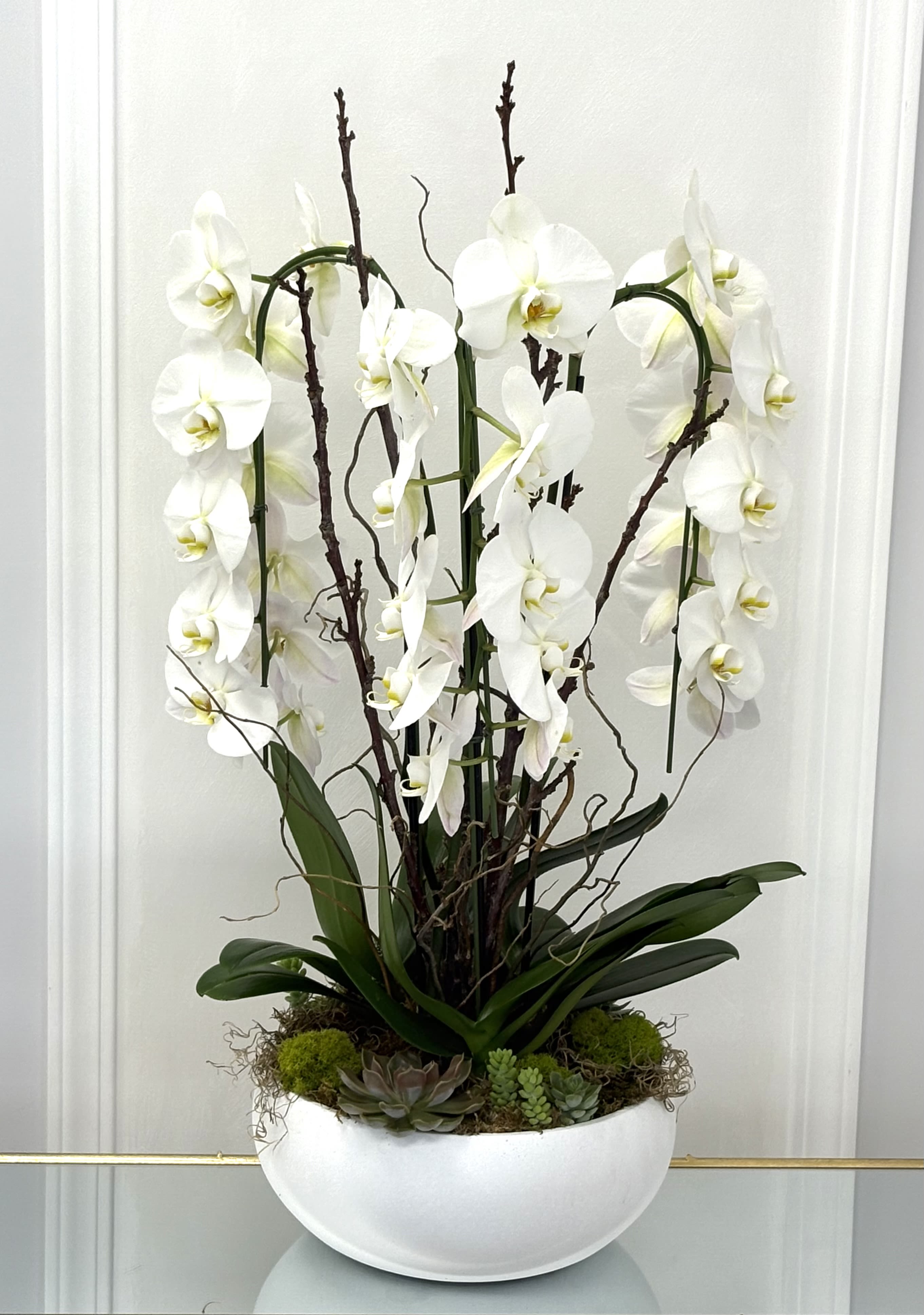 Whispering Grace By VLM Flowers - An exquisite arrangement of cascading white Phalaenopsis orchids, artfully paired with delicate curly willow branches and grounded by a base of moss, succulents, and natural textures. Displayed in a modern white ceramic bowl, this design radiates purity, peace, and timeless elegance. Ideal for sympathy gestures, serene home décor, or celebrating life’s most meaningful moments with sophistication and calm.