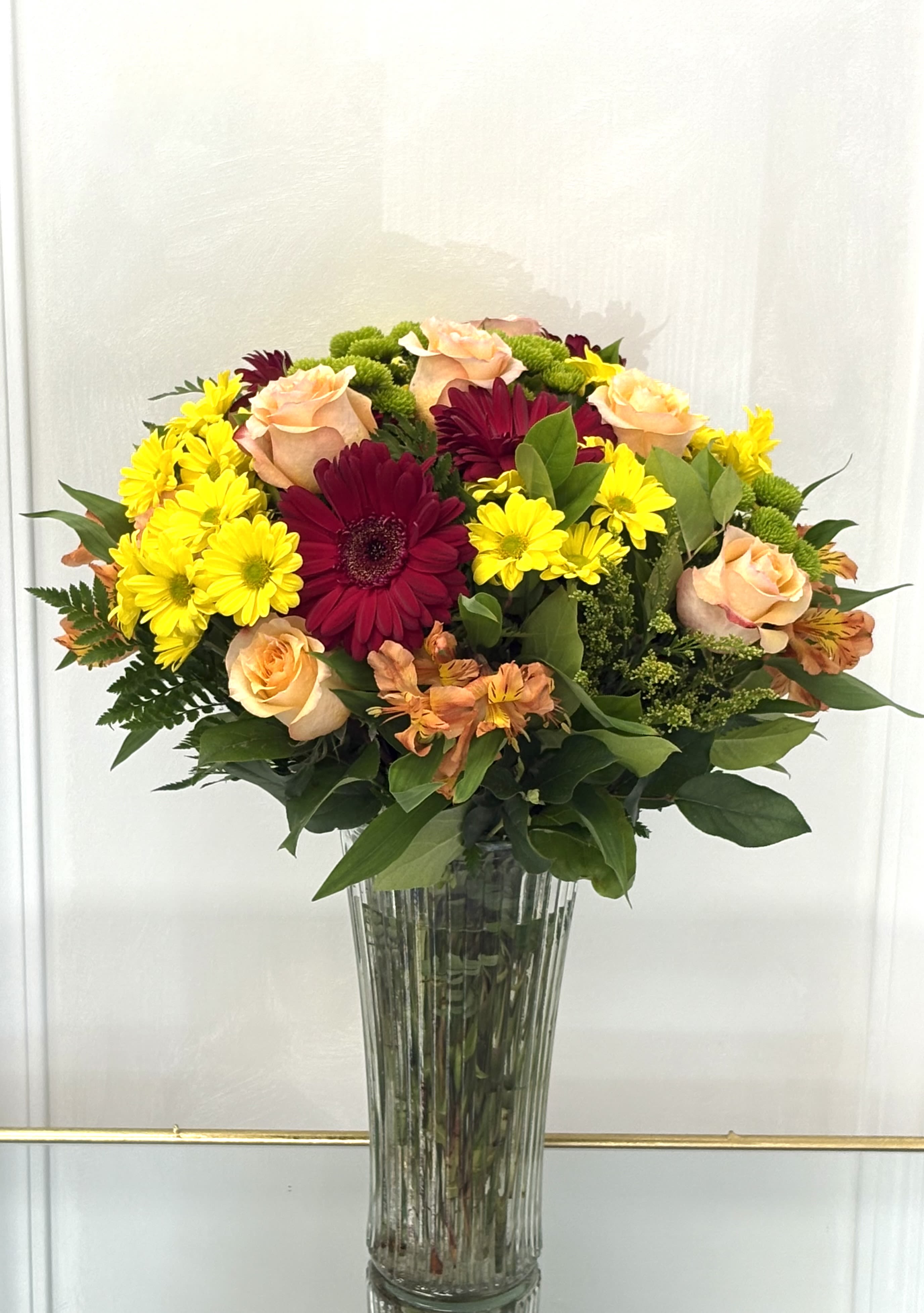  Sunset Celebration By VLM Flowers - Inspired by the glow of a summer sunset, this lively arrangement mixes soft peach roses, deep red gerberas, golden daisies, and playful greens. Presented in a clear vase, it radiates happiness, energy, and heartfelt cheer.  Perfect For: Birthdays, congratulations, or get-well wishes.