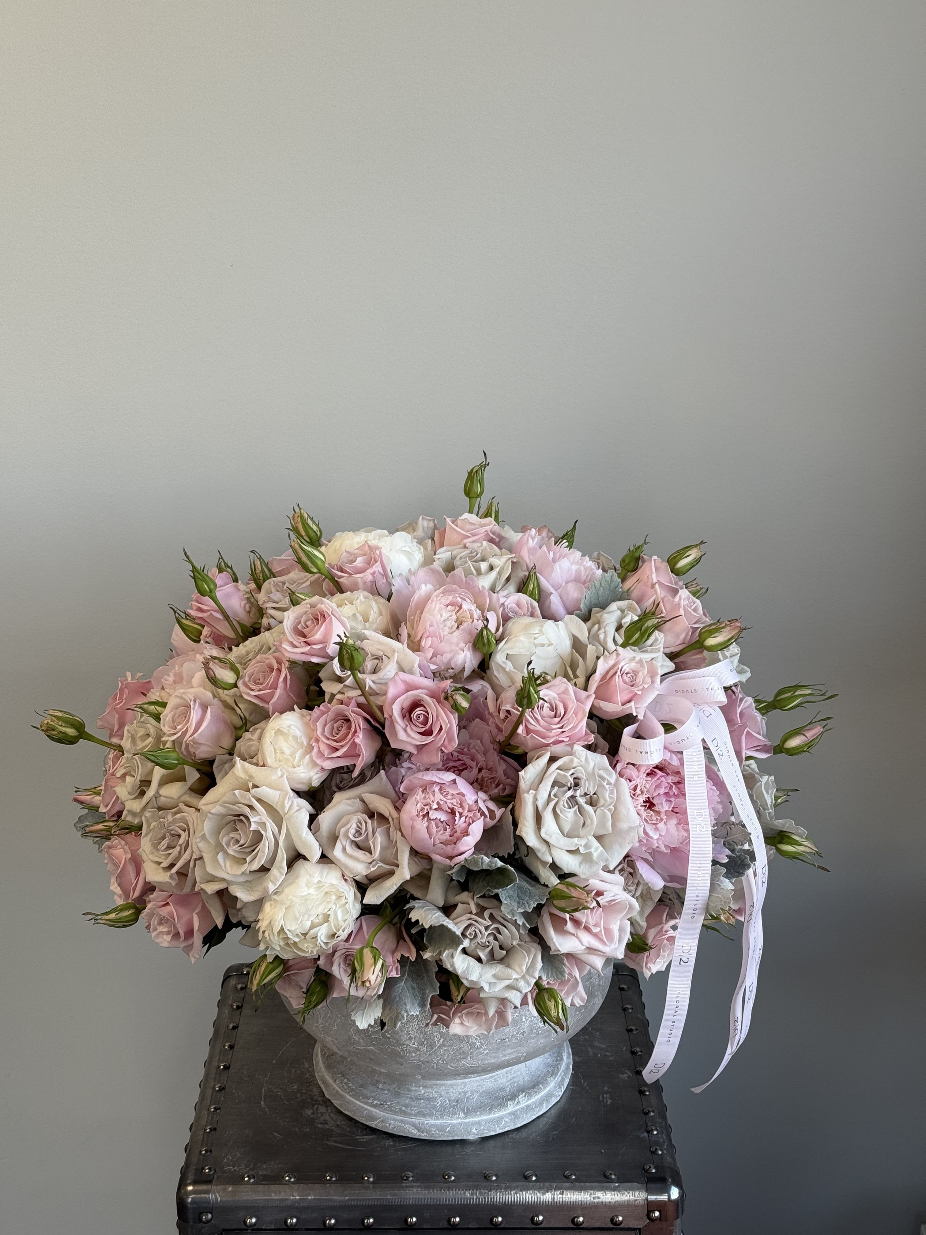 Icelynn - A romantic and graceful arrangement in soft blush, ivory, and dusty rose tones, featuring peonies, spray roses, and lisianthus buds. Artfully arranged in a textured white pedestal vase and adorned with delicate pale pink D|2 ribbons—ideal for weddings, anniversaries, or elegant gift-giving.