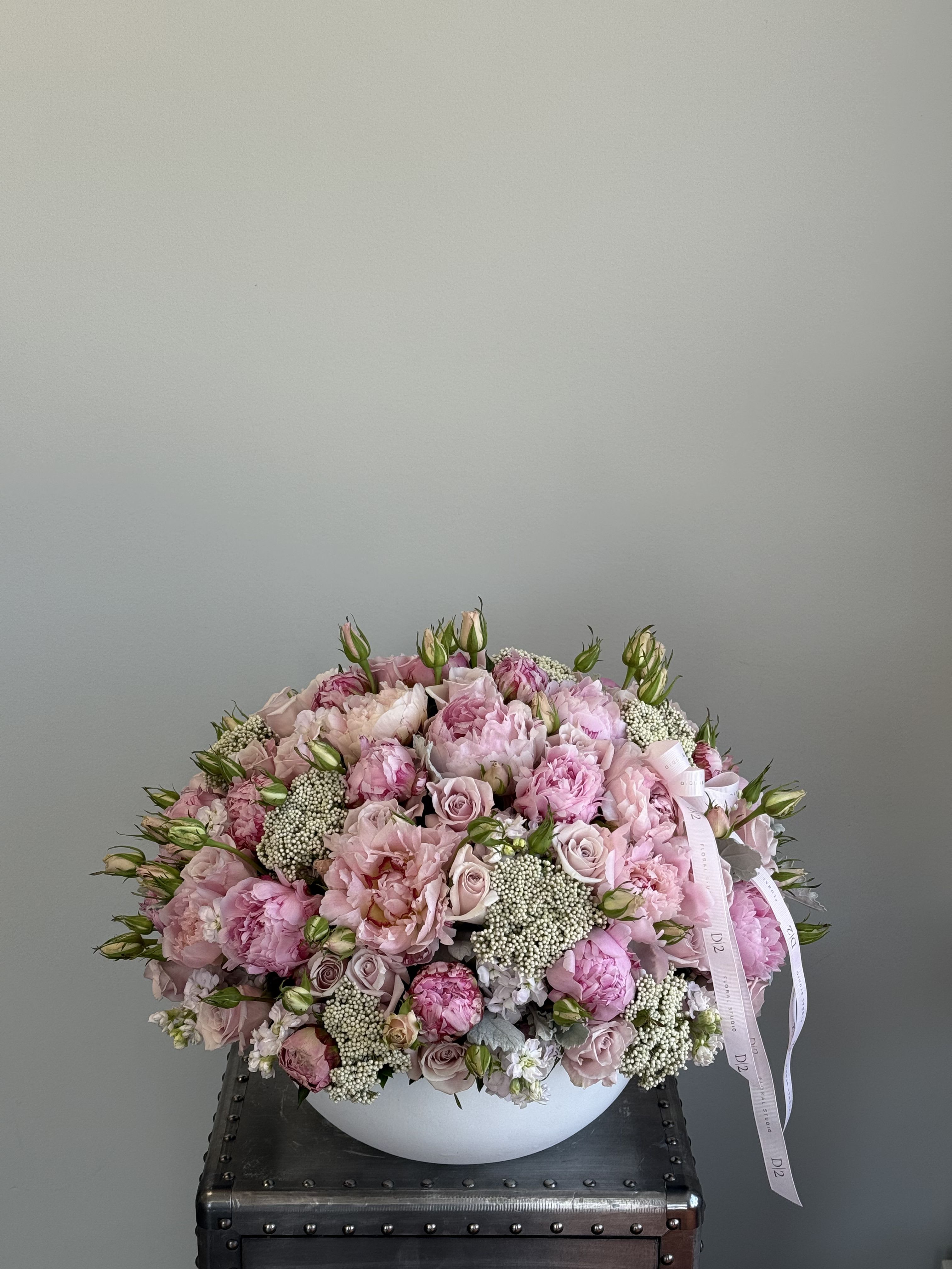 Hallie - A charming and lush arrangement featuring soft pink peonies, blush roses, lisianthus buds, and clusters of delicate white filler flowers. Artfully arranged in a round white ceramic vase and adorned with pastel pink D|2 ribbons—perfect for romantic gestures, birthdays, or welcoming a new baby girl.