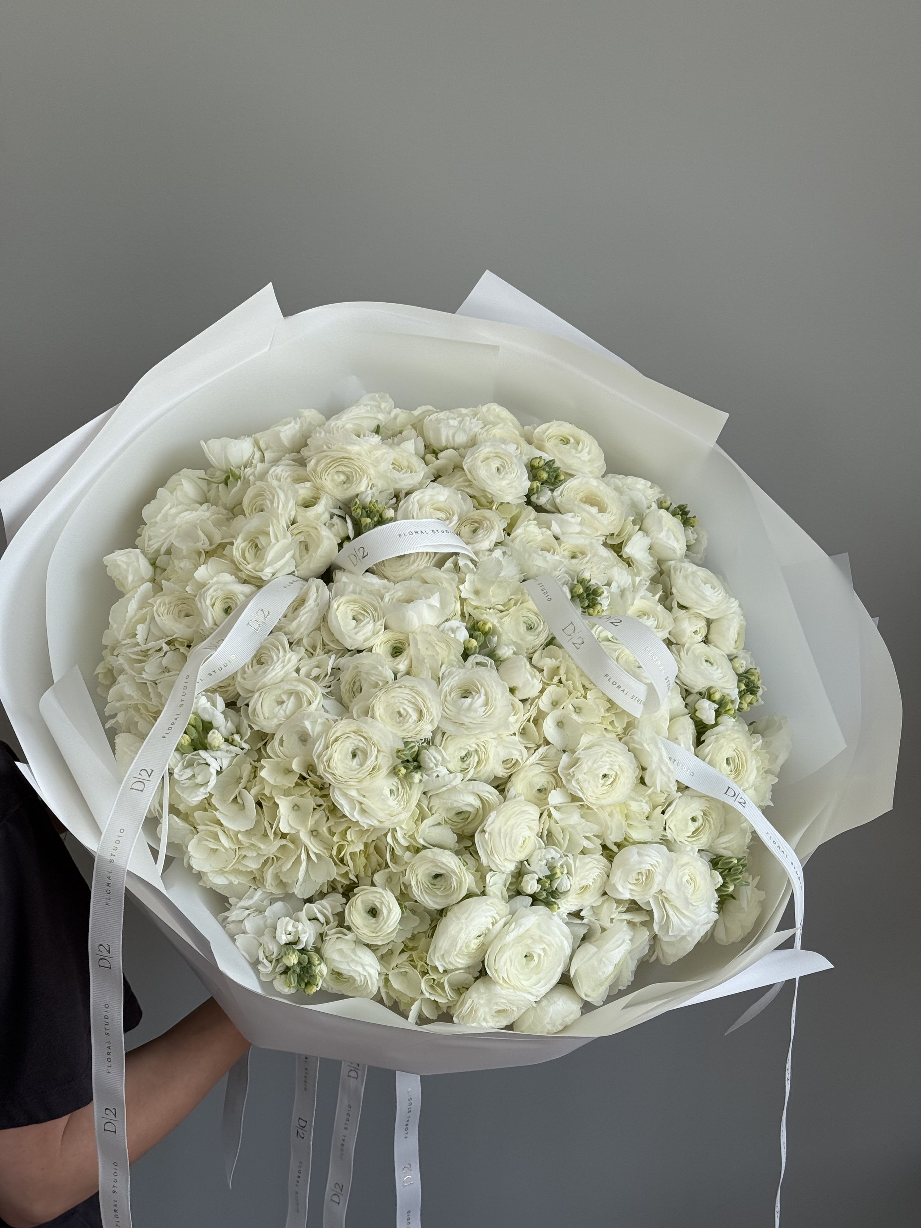 Ruth - A breathtaking bouquet composed entirely of pristine white ranunculus and delicate filler blooms, wrapped in layers of soft white paper and tied with elegant white D|2 ribbons. A timeless and graceful arrangement, perfect for weddings, sympathy, or sophisticated celebrations.