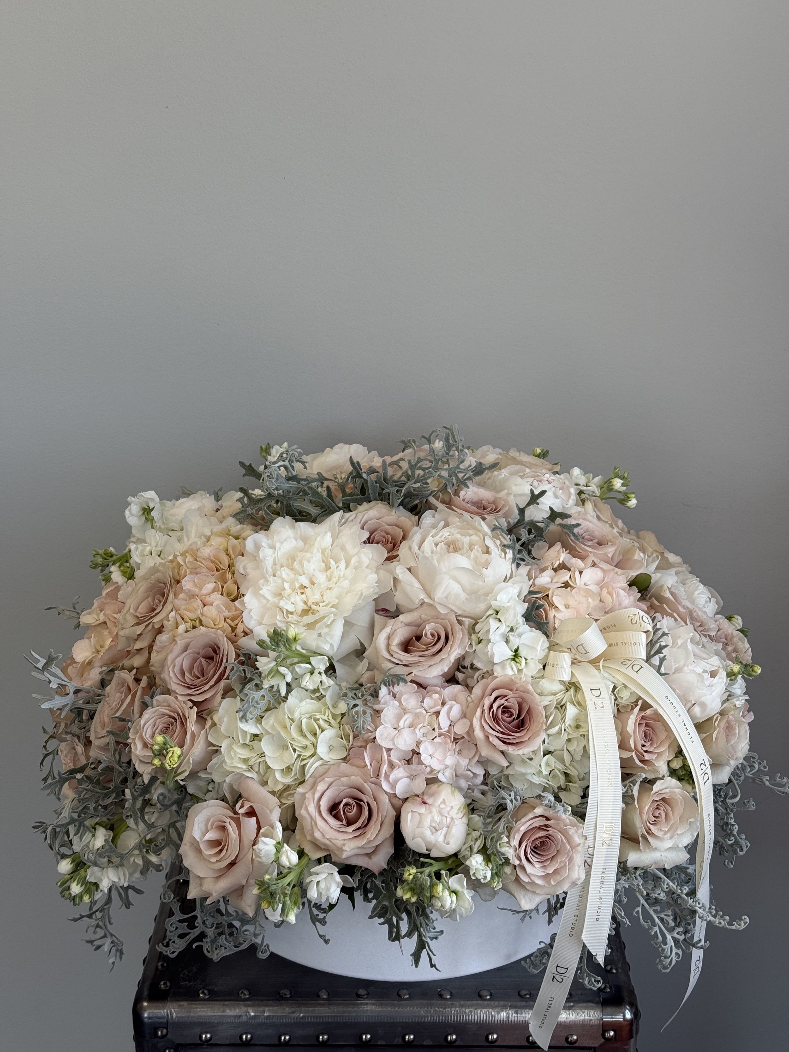Tatum - A luxurious and soft-toned arrangement featuring blush and ivory roses, hydrangeas, peonies, and lisianthus, accented with silvery dusty miller foliage. Presented in a sleek white ceramic bowl and finished with a cream D|2 ribbon—perfect for elegant weddings, sympathy tributes, or timeless celebrations.
