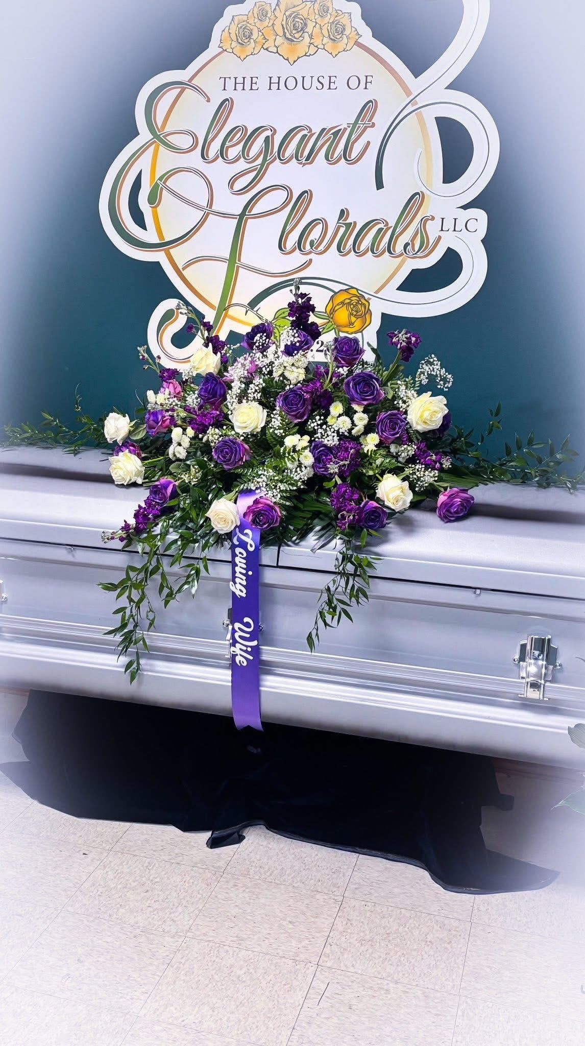 Eternal Grace – Purple &amp; Ivory Rose Casket Spray - A timeless and dignified tribute, our Eternal Grace Casket Spray blends rich hues of purple and soft ivory to honor a life beautifully lived. Designed with cascading elegance, this full casket spray features premium lavender and royal purple roses, creamy ivory roses, delicate baby’s breath, and a lush bed of greenery.  Adorned with a satin ribbon that reads “Loving Wife,” this arrangement expresses both sorrow and reverence with unmatched beauty—perfect for celebrating the memory of a cherished woman who brought grace and love to every moment.  Details:  Flowers: Purple roses, ivory roses, white daisies, baby’s breath, mixed greenery  Size: Full casket length (approx. 36–48” wide)  Includes: “Loving Wife” tribute ribbon (custom text available upon request)  Ideal for: Funeral services, celebration of life, homegoing ceremonies  
