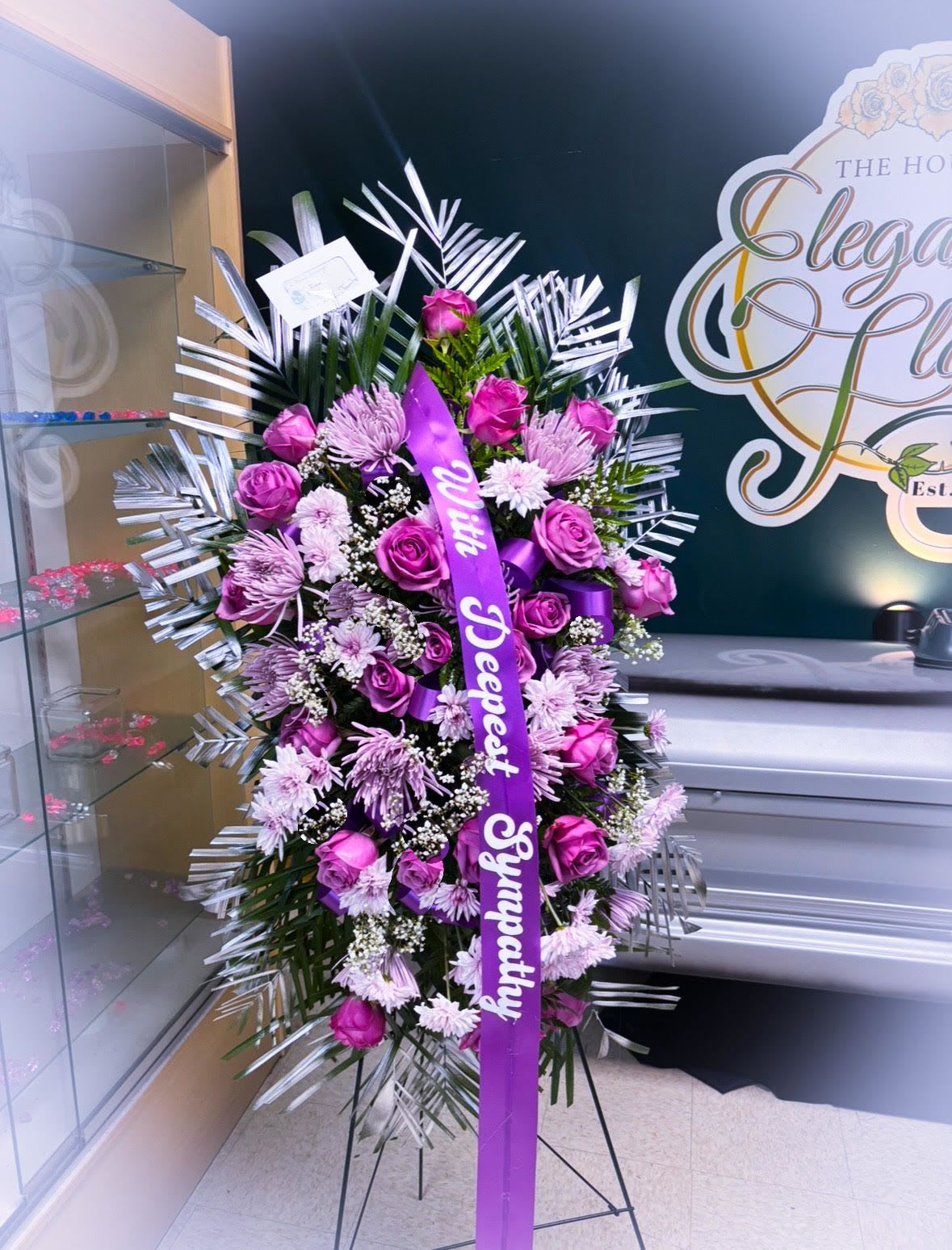 Royal Lavender Tribute – Standing Sympathy Spray - Graceful, regal, and deeply heartfelt, our Royal Lavender Tribute Standing Spray is a striking display of remembrance designed to honor a beautiful life. This tall, elegant arrangement features vibrant lavender roses, soft purple spider mums, delicate baby’s breath, and lush palm greenery—framed with shimmering silver foliage for a modern, dignified touch.  Adorned with a satin ribbon that reads “With Deepest Sympathy”, this standing spray offers a powerful yet comforting presence at funeral services, memorials, and homegoing celebrations.    Details:  Flowers: Lavender roses, purple spider mums, baby’s breath, silver accents, palm greenery  Display: Mounted on a standing easel (included)  Size: Approx. 36–48” tall  Ribbon Message: “With Deepest Sympathy” (customizable)  Style: Elegant, modern, and serene  Ideal for: Funerals, homegoings, celebration of life services