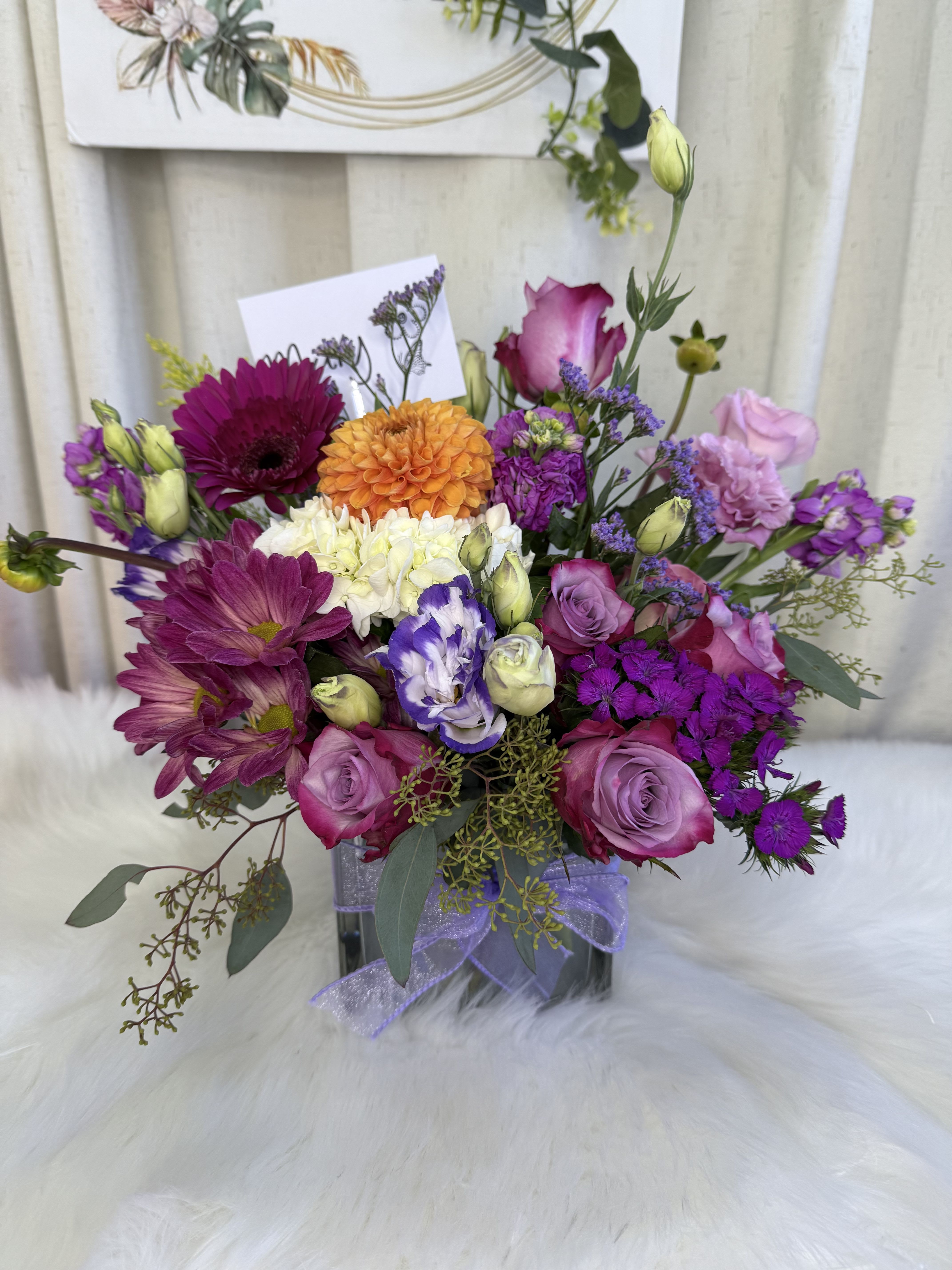 Color Charm” Floral Arrangement  - Color Charm, Floral Arrangement A vibrant composition blending the elegance of lavender roses, the freshness of fuchsia gerberas, the brightness of an orange dahlia, and the delicate beauty of lisianthus in cream and purple tones. Designed in a modern glass vase with a decorative ribbon, this arrangement is perfect for brightening any special occasion and expressing emotions with style and freshness.