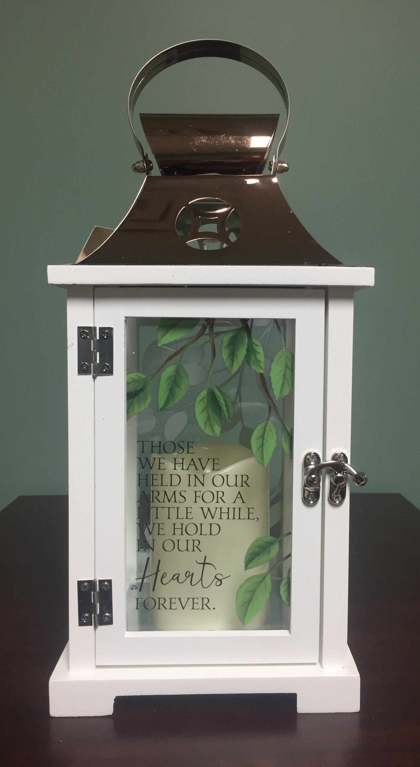 You're In Our Hearts Forever Lantern - This beautiful sympathy themed lantern is a thoughtful lasting tribute suitable for home delivery or funeral home display. It contains a flameless LED candle and is made from quality wood with stainless steel copper finish top. Equipped with a built-in automatic timer and 3 AAA batteries. Size:13 1/2inch H x 6 1/4 inch W x 6 1/4 inch D. On the front glass panel the lantern reads: THOSE WE HAVE HELD IN OUR ARMS FOR A LITTLE WHILE, WE HOLD IN OUR HEARTS FOREVER.  The saying is surrounded by beautiful green leaves on flowing branches.       