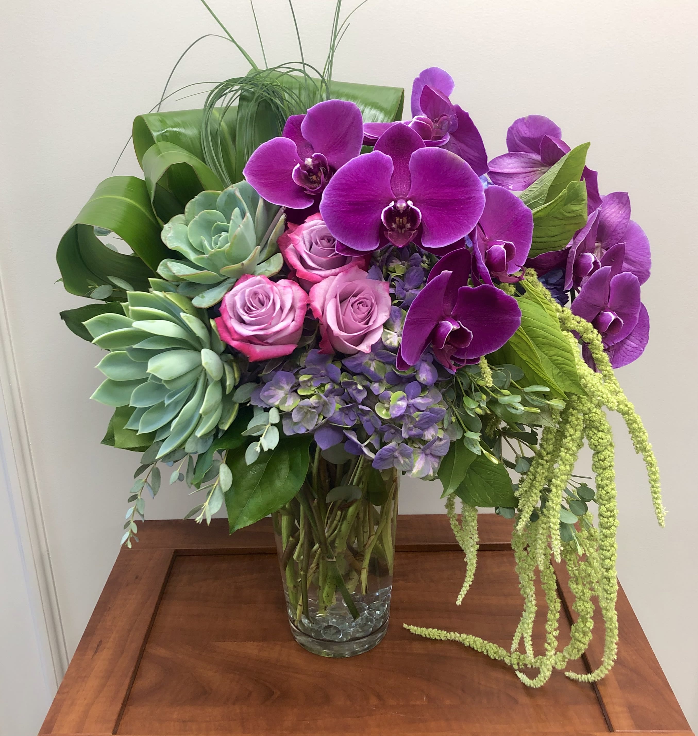 Exquisite Elegance Bouquet - A sculpture of blooms. Elevate any occasion with this exquisite masterpiece of elegant orchids, roses and graphic succulents, dripping with amaranthus and arranged in an oversized glass jug. This eye-catching bouquet features purple hydrangea, lavender roses, lavender phalaenopsis spray orchids, hanging green amaranthus, bear grass, green ti leaves, and large green echeveria succulents. Delivered in a clear milk jug vase. Orientation: All-Around Design in photo is showing the Premium version. We create this design in 3 different price points. 