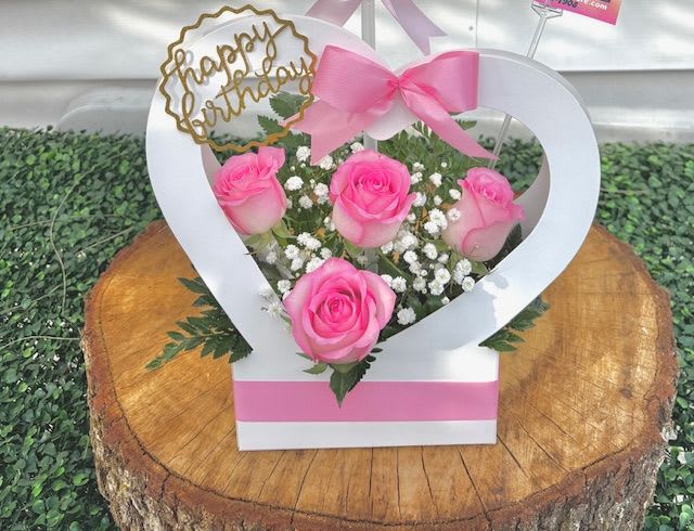 Whispers of Love - A graceful heart-shaped basket, sitting in pristine white, symbolizing purity, love, and sincerity. Nestled within its elegant curves are fresh pink roses in delicate gradients—from blush to rosewood—each bloom unfolding softly like a whisper of affection. Their velvety petals are arranged with care, fanning outward in a harmonious blend of color and form.  Atop this tender composition sits a beautiful bow, crafted from luminous satin ribbon in a gentle pink hue. Its tails drape gracefully over the roses, adding a touch of sophistication and charm, like the final note in a love song. The basket may also include accents like baby's breath or greens to heighten its texture and elegance.