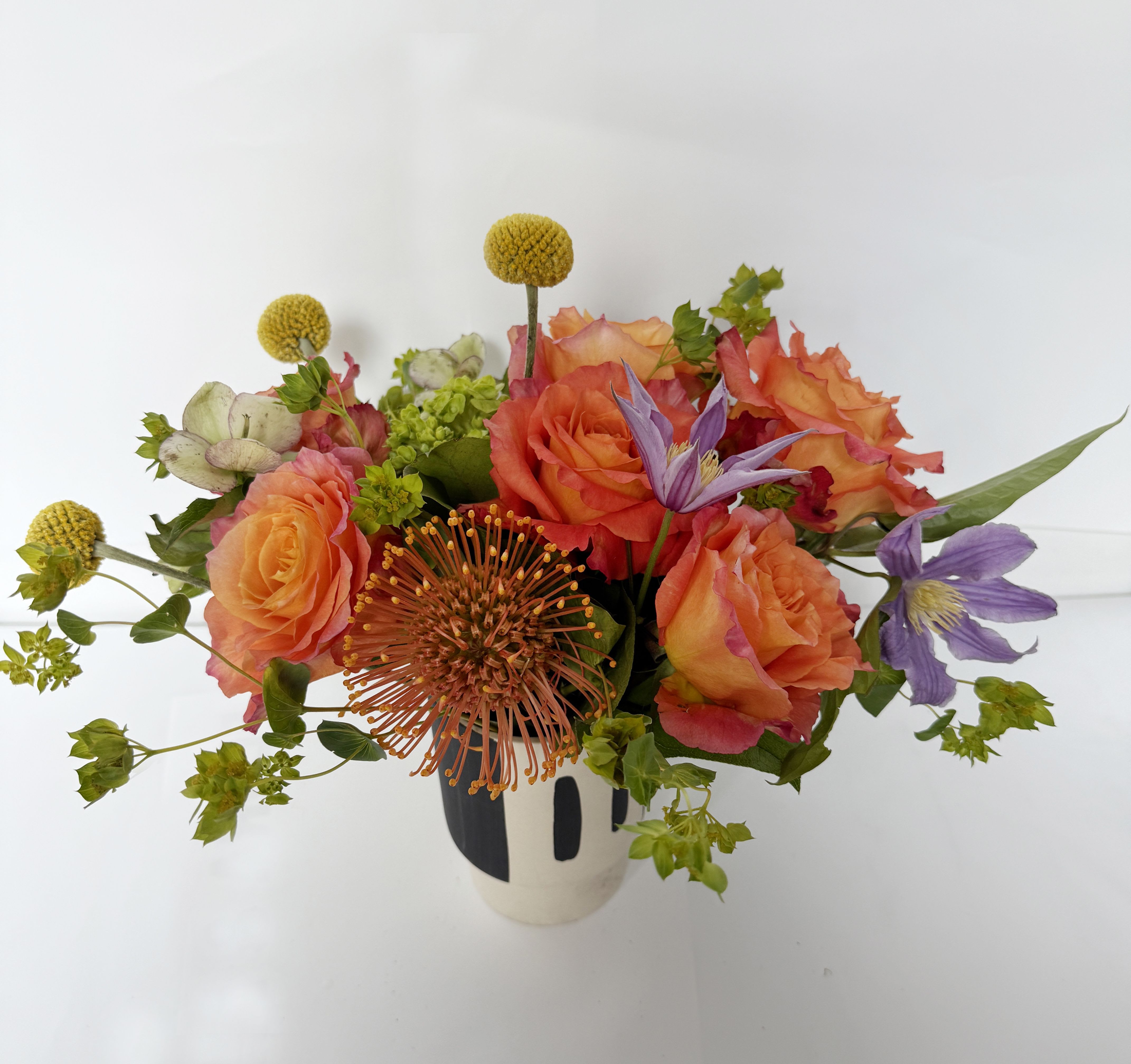 Trabar - Bold, fiery, and full of flair — Trabar is a dynamic mix of color and form that makes a statement in any space. This contemporary arrangement features ruffled bi-color orange roses, whimsical billy balls (Craspedia), striking purple clematis, and sculptural pincushion protea, all artfully arranged in a modern black-and-white ceramic vase.  A perfect piece for the creative soul, this design adds instant energy and personality to desks, dining tables, or gifting moments.  Details: 	•	Featuring roses, clematis, protea, billy balls, hellebores, and seasonal greenery 	•	Approx. size: 9-10” H x 7-8” W 	•	Designed in a bold black-and-white ceramic vase 	•	Includes a free personalized message card  Care Instructions: Display in a cool, well-lit spot out of direct sun. Change water daily and trim stems slightly every 2–3 days to prolong freshness.