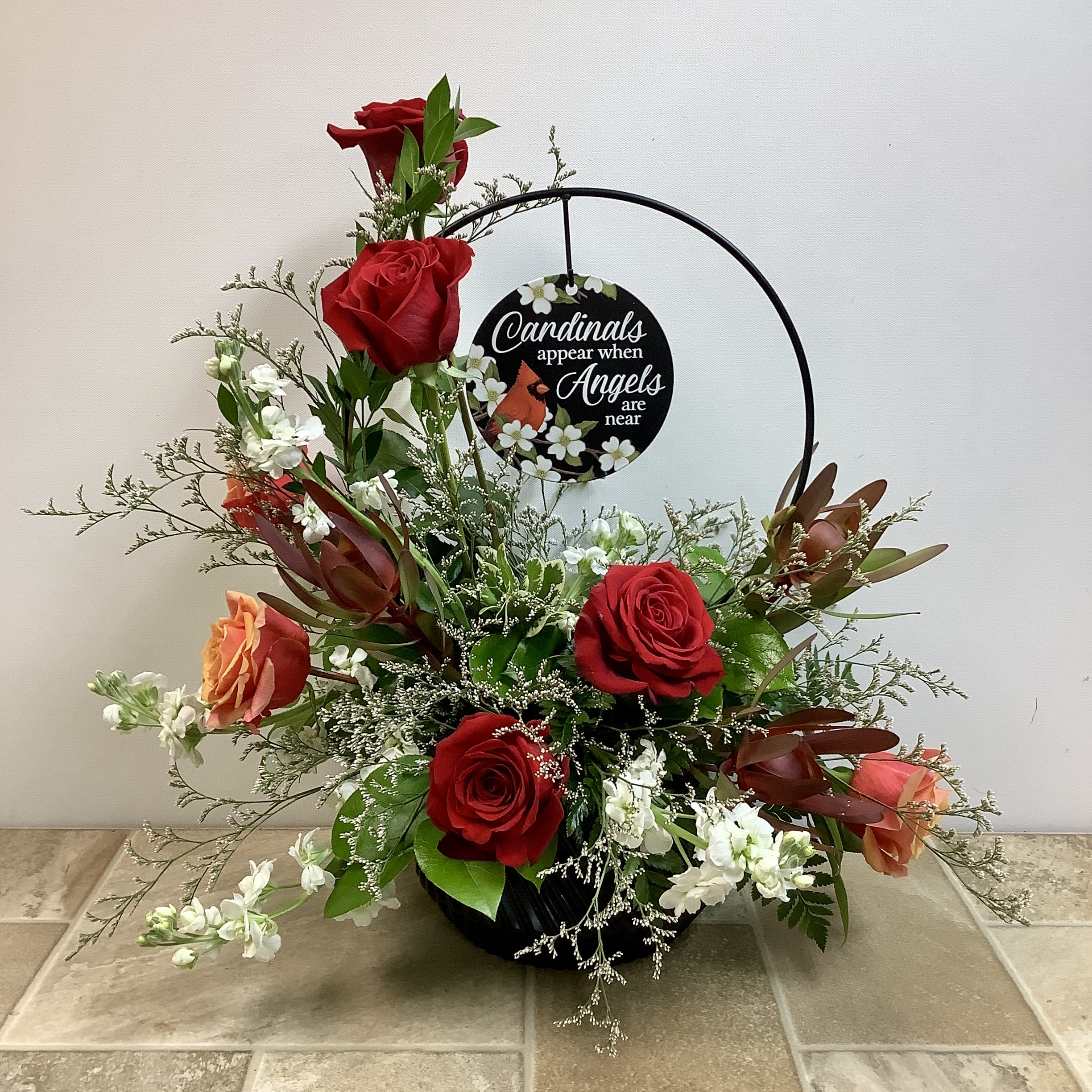 Signs of Love Signs of Love - This elegant tribute piece features a mix of vibrant and earthy tones, anchored by deep red roses that symbolize love and remembrance. Peach-toned Cherry Brandy roses contribute warmth, burgundy leukadendron add a modern touch, and white stock flowers represent purity and peace. Accents of soft greenery and wispy white limon provide texture and airiness. The standout element is a keepsake circular metal hoop displaying a heartfelt sign that reads: "Cardinals appear when Angels are near." Approximate size: 21" wide x 22" tall Primary flowers: rose, conebush