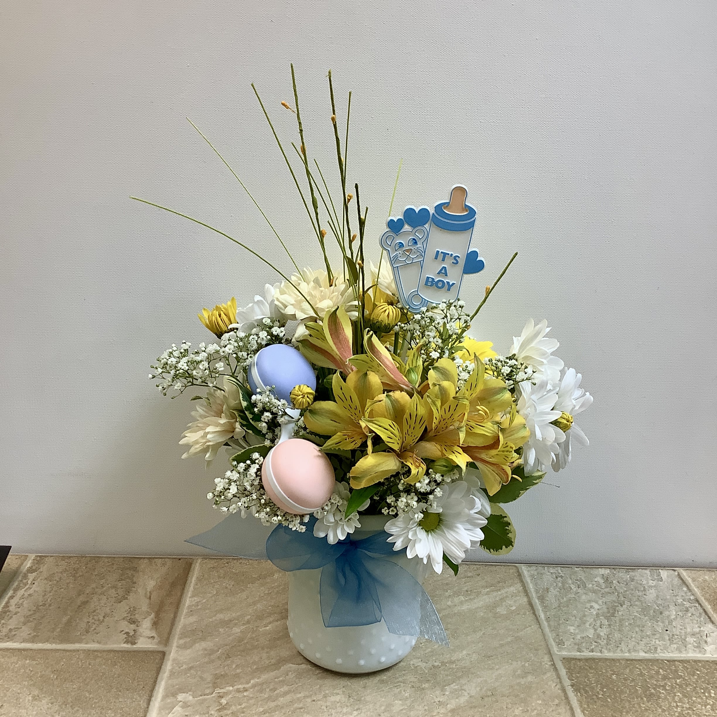 Welcome Baby Boy - Send a happy message of welcome and congratulations with this sweet arrangement in a white, hobnail vase with a blue bow. It features a peach cushion mum, white and yellow daisies, yellow alstroemeria, and baby's breath. The bouquet is accented with wispy stems and yellow silk sprigs for added height and texture.  A baby bottle sign announces &quot;It's a boy&quot; and a blue and pink rattle completes the celebratory, baby-related theme.  Approximate size: 11&quot; wide x 15&quot; tall  Primary flowers: daisy, chrysanthemum, mums, lily