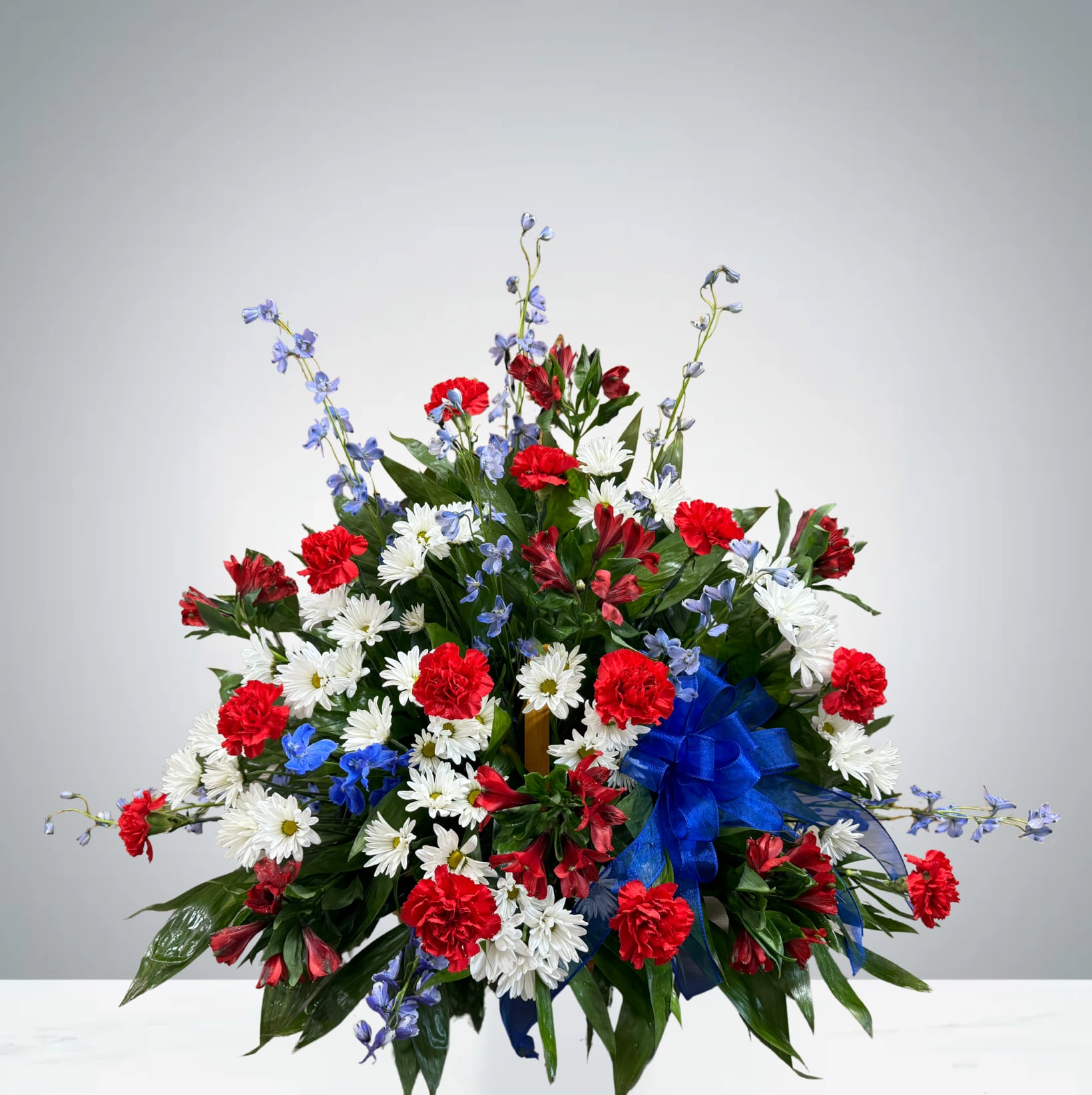Harmony in Red, White & Blue Harmony in Red, White & Blue - Celebrate the spirit of patriotism and beauty with this vibrant flower basket arrangement. Featuring striking blue delphinium, bold red carnations, elegant white daisies, and rich red alstroemeria, this design bursts with classic American charm. Nestled in a beautiful woven basket and adorned with large, eye catching bow, it’s accented with fresh greenery to bring the entire piece to life. Perfect for funerals, holiday, tributes, or simply showing your pride, this arrangement is heartfelt display of color, honor, and natural grace.