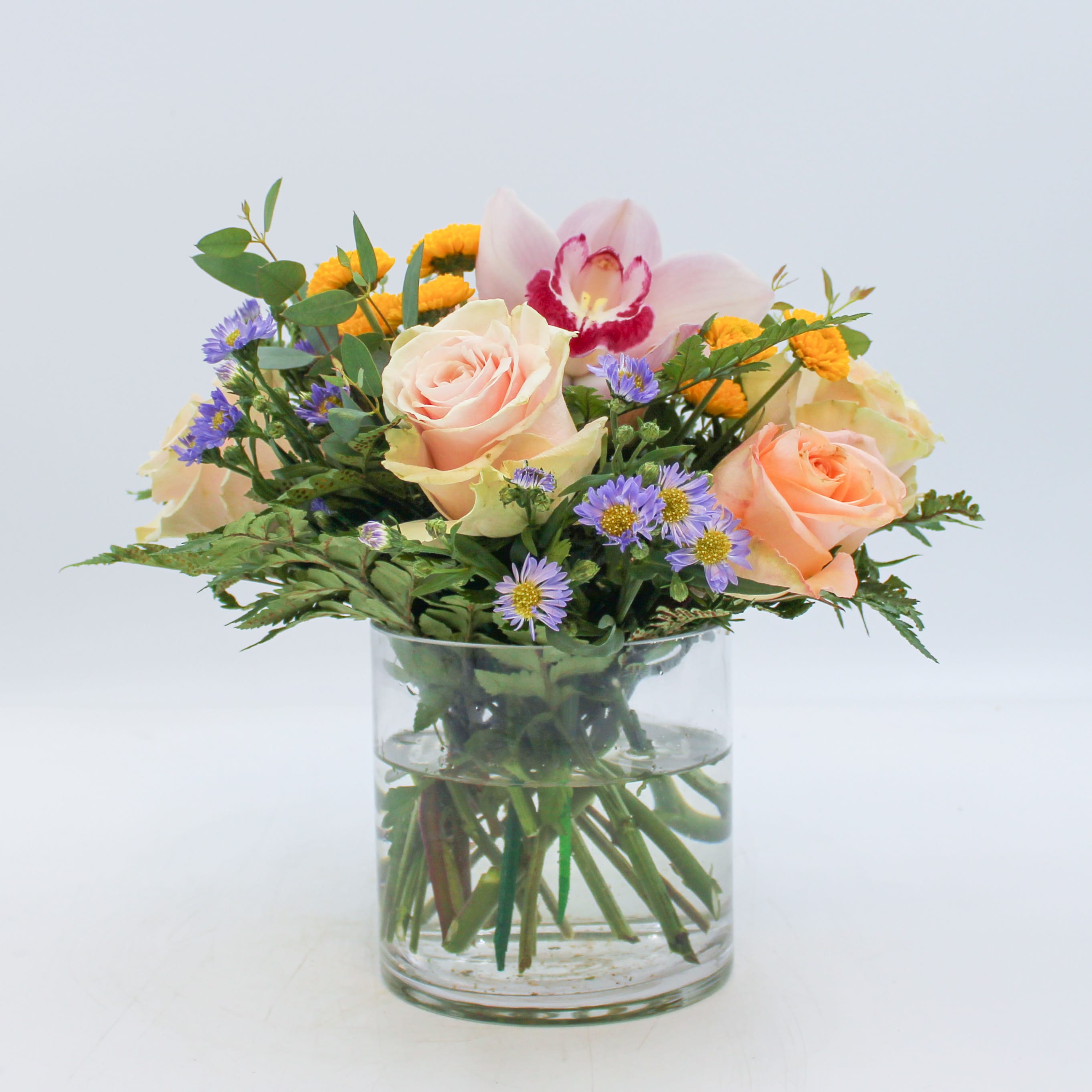 Poppy - Poppy features a Pink Cymbidium Orchid bloom, Blush Pink Roses, Peach Roses, Yellow Button Mums, Purple and Mixed foliage arranged in a clear glass vase. 