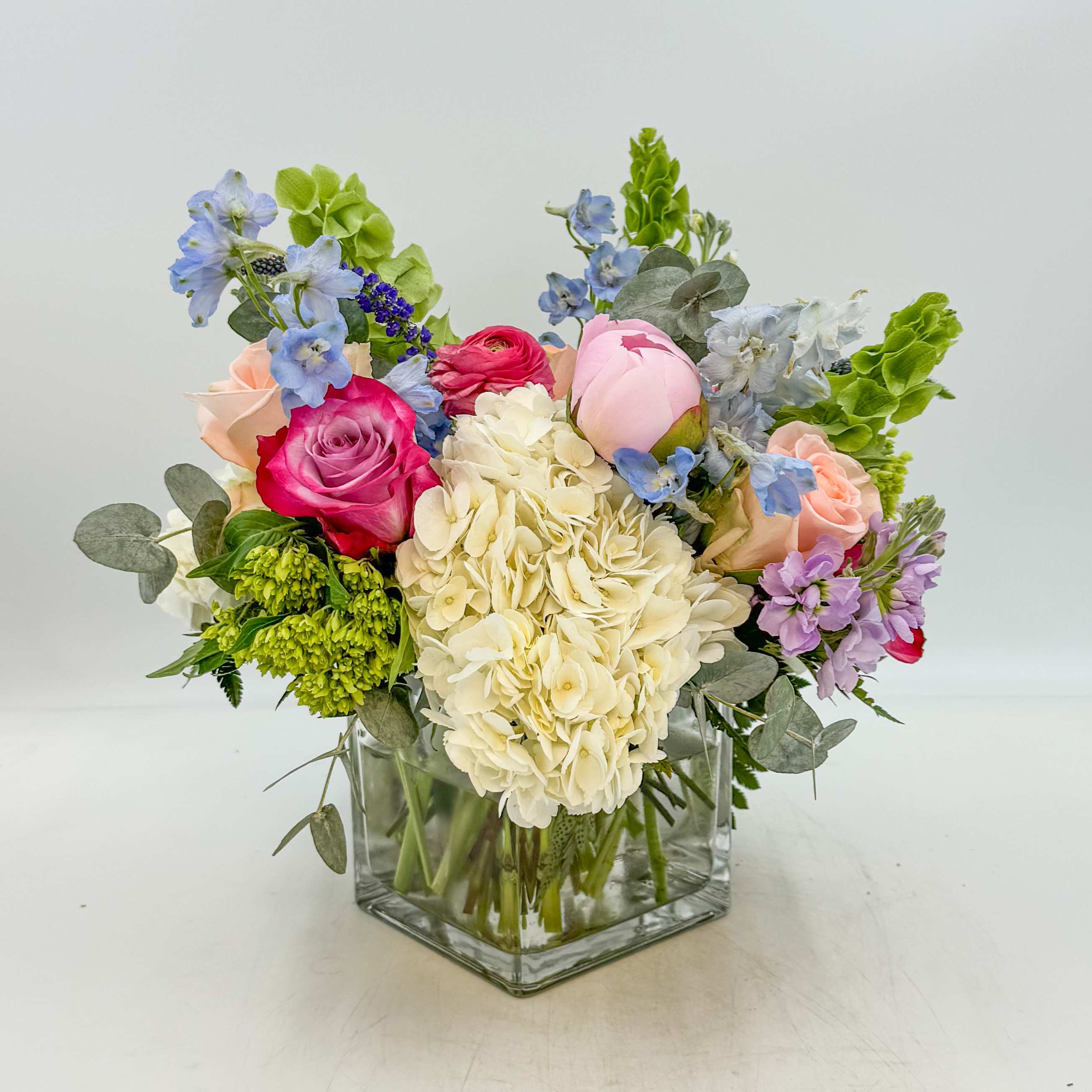 Summer Bliss - Summer Bliss features White Hydrangea, Mini Green Hydrangea, Pink Peonies, Peach Roses, Deep Purple Roses, Lavender Stock, Blue Delphinium, Bells of Ireland, Hot Pink Ranunculus, Purple Veronica with Eucalyptus. Measurements: The Standard Summer Bliss is designed in a 5 inch glass square vase; the Deluxe Summer Bliss is designed in a 6 inch glass square vase; and the Premium Summer Bliss is designed in an 8 inch glass square vase.