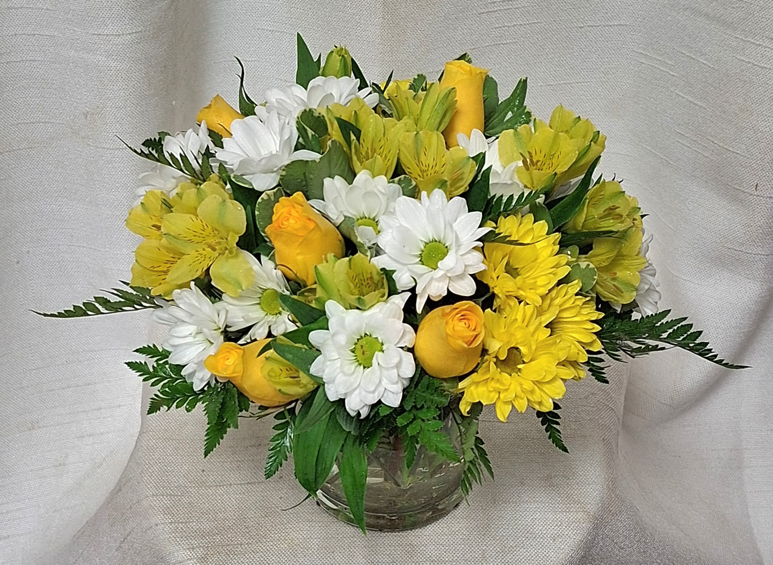 Mellow Yellow (BB) - Introducing our simple yet modern floral arrangement that exudes beauty and elegance. This arrangement features a stunning combination of yellow roses, yellow alstroemeria, white daisies, and white button poms, artfully arranged in a clear glass vase.