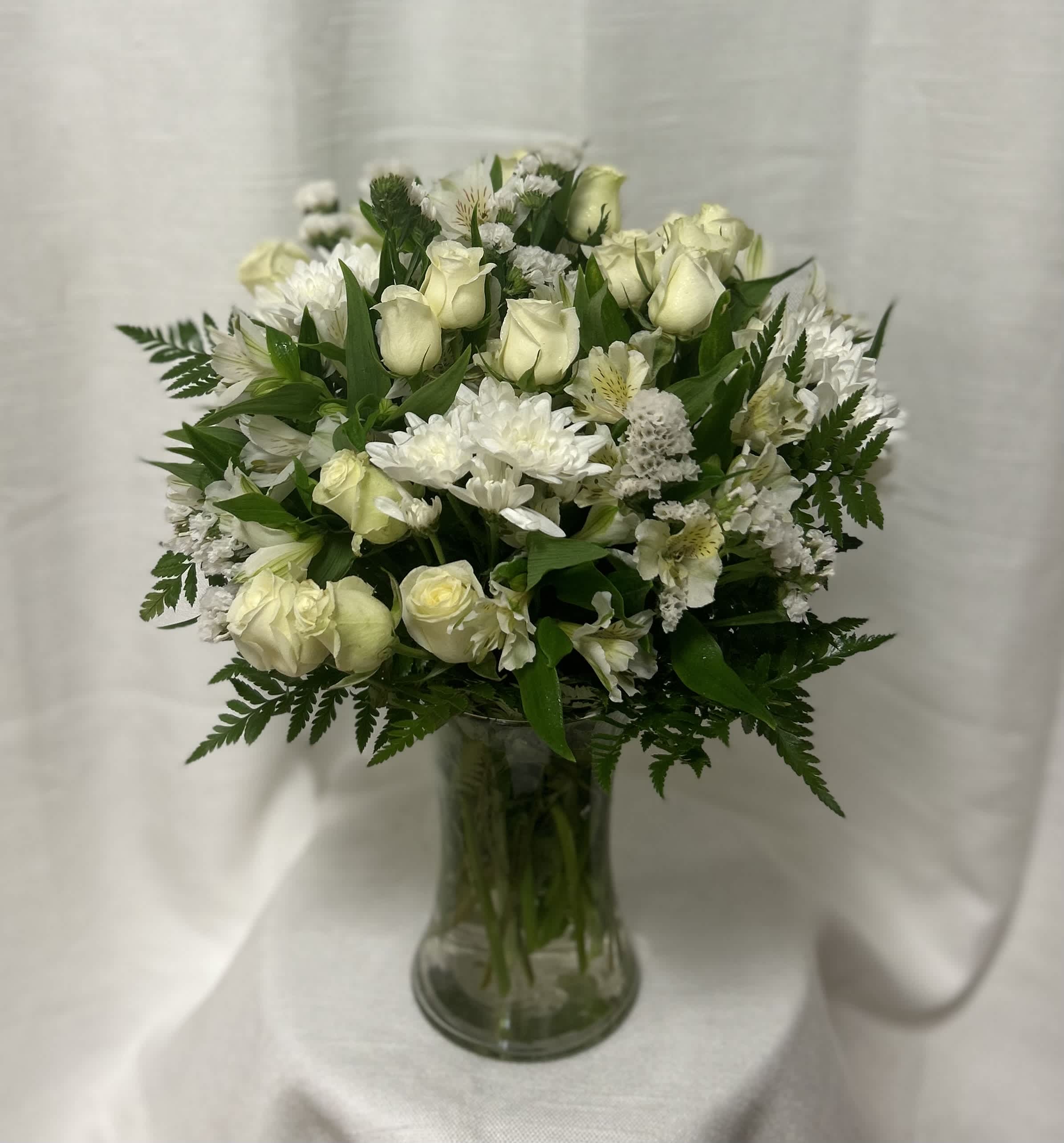 Forever Friendship - During this difficult time, we offer a serene and elegant funeral flower arrangement that is a beautiful tribute to honor and celebrate the cherished memories of your loved one. This arrangement features a beautiful combination of white roses, white alstroemeria, and premium leather leaf, carefully arranged in a clear glass vase to create a serene and peaceful atmosphere.  