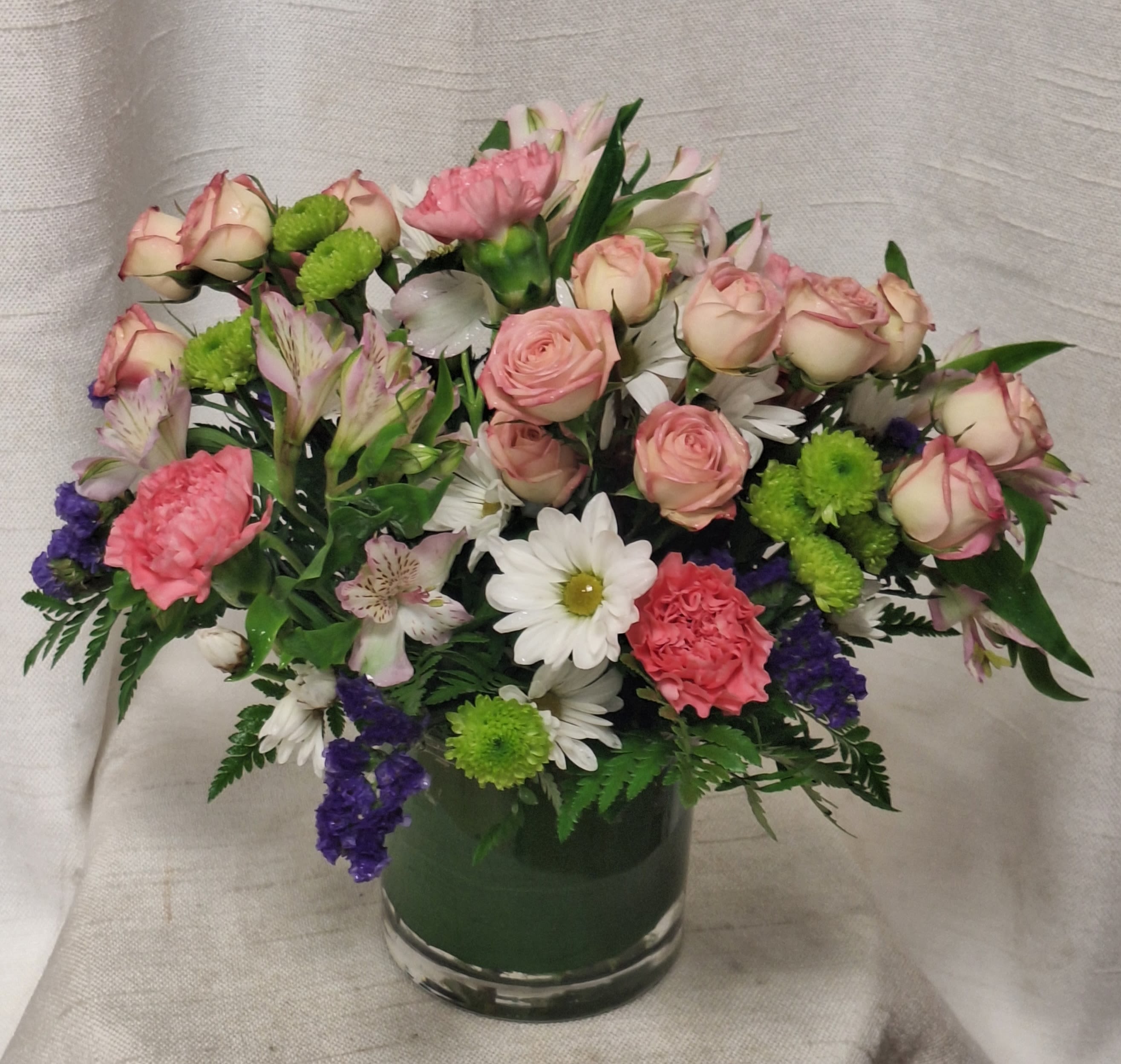 Berry Bliss (BB) - Introducing our Berry Bliss Floral Arrangement, a vibrant and charming display of nature's beauty. This delightful arrangement features a mix of purple statice, green button poms, pink spray roses, white daisies, purple alstroemeria, and hot pink carnations, all lovingly arranged in a clear glass vase.
