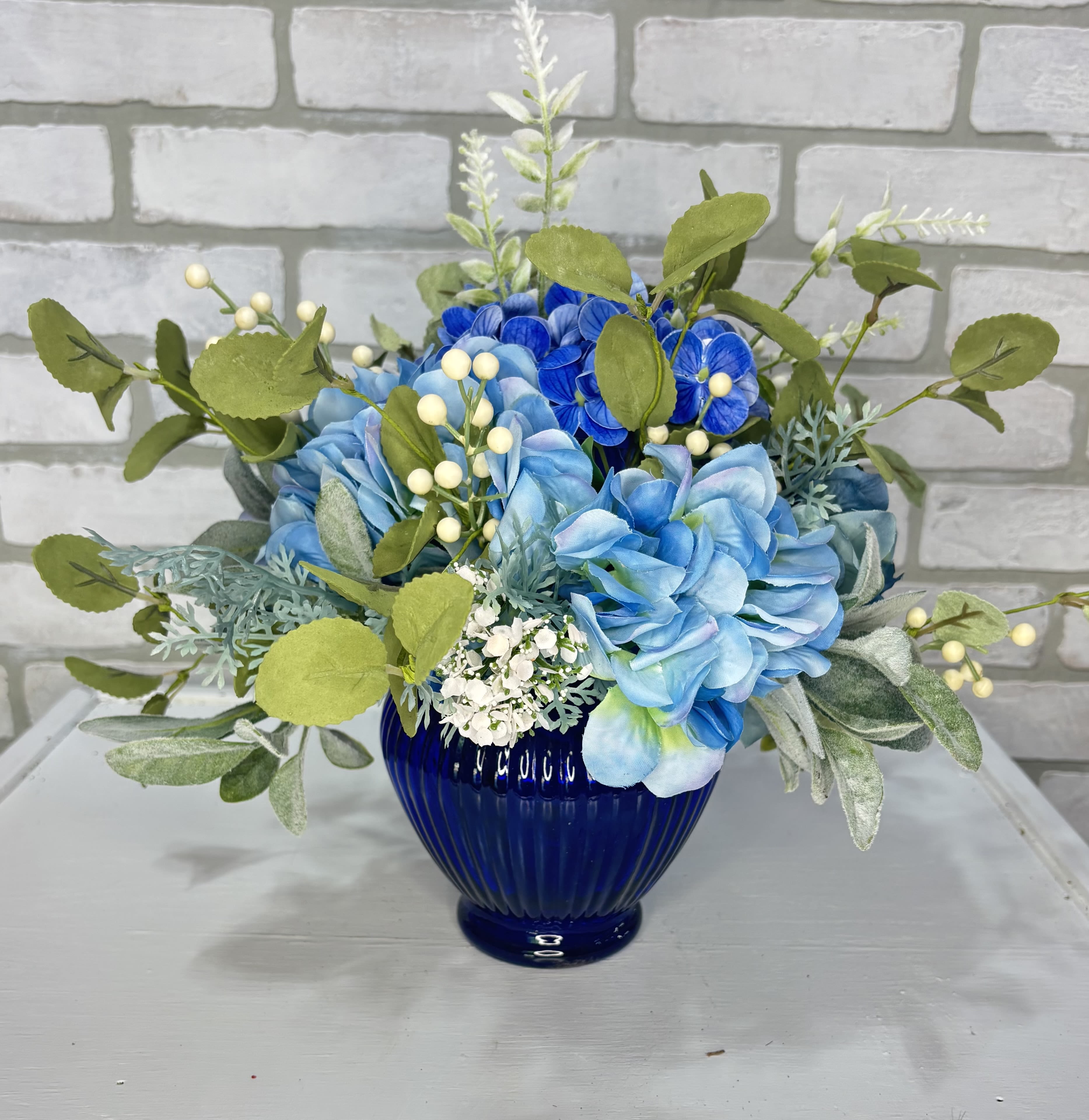 Blue Artificial Silk Arrangement Blue Artificial Silk Arrangement - A blue vase arranged with artificial blue hydrangea, baby's breath, hypericum berries, and mixed greens.