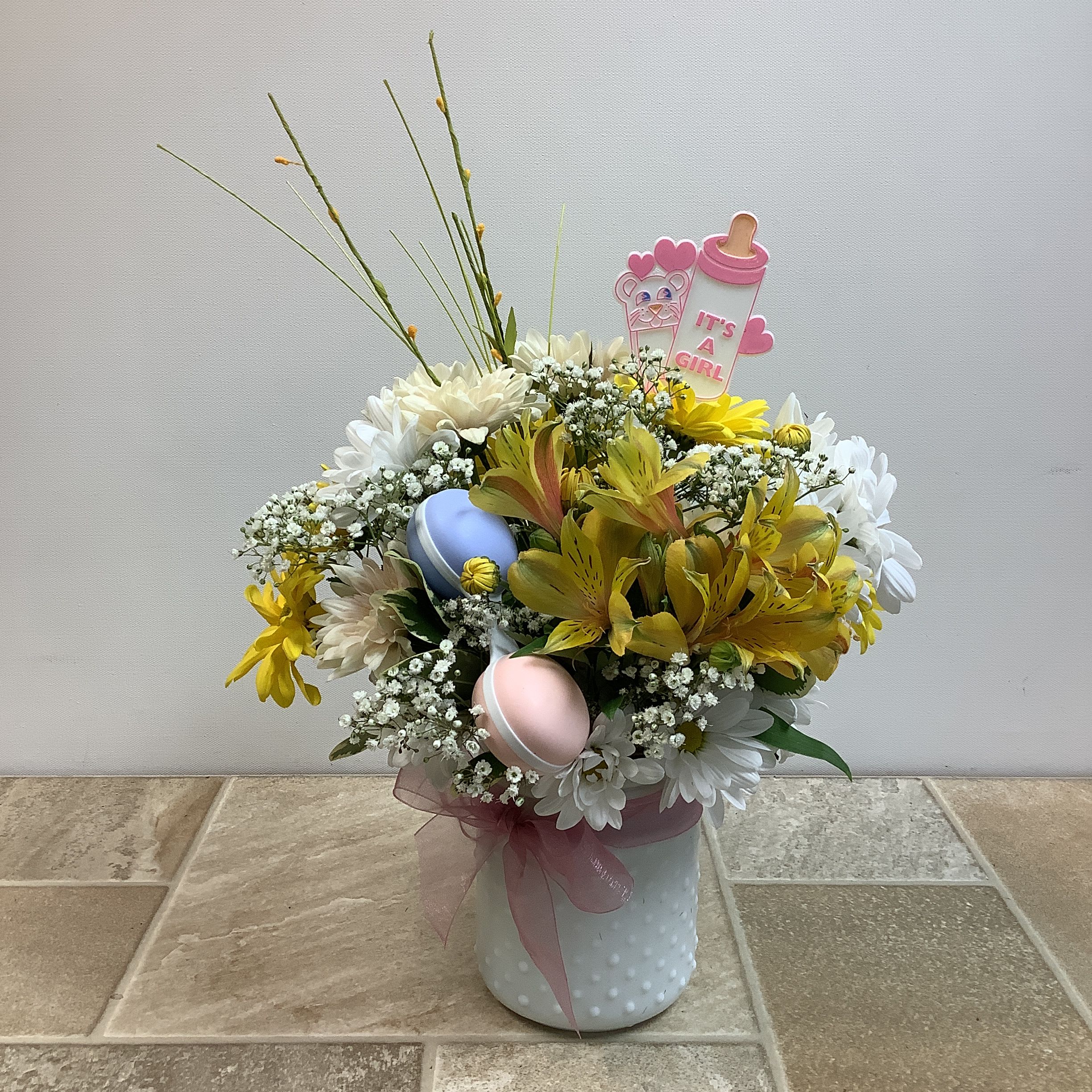 Welcome Baby Girl Welcome Baby Girl - Send a happy message of welcome and congratulations with this sweet arrangement in a white, hobnail vase with a pink bow. It features a peach cushion mum, white and yellow daisies, yellow alstroemeria, and baby's breath. The bouquet is accented with wispy stems and yellow silk sprigs for added height and texture. A baby bottle sign announces "It's a girl", and two macaron-shaped decorations, one light blue and one light pink, complete the celebratory, baby-related theme. Approximate size: 11" wide x 15" tall Primary flowers: daisy, chrysanthemum, mums, lily