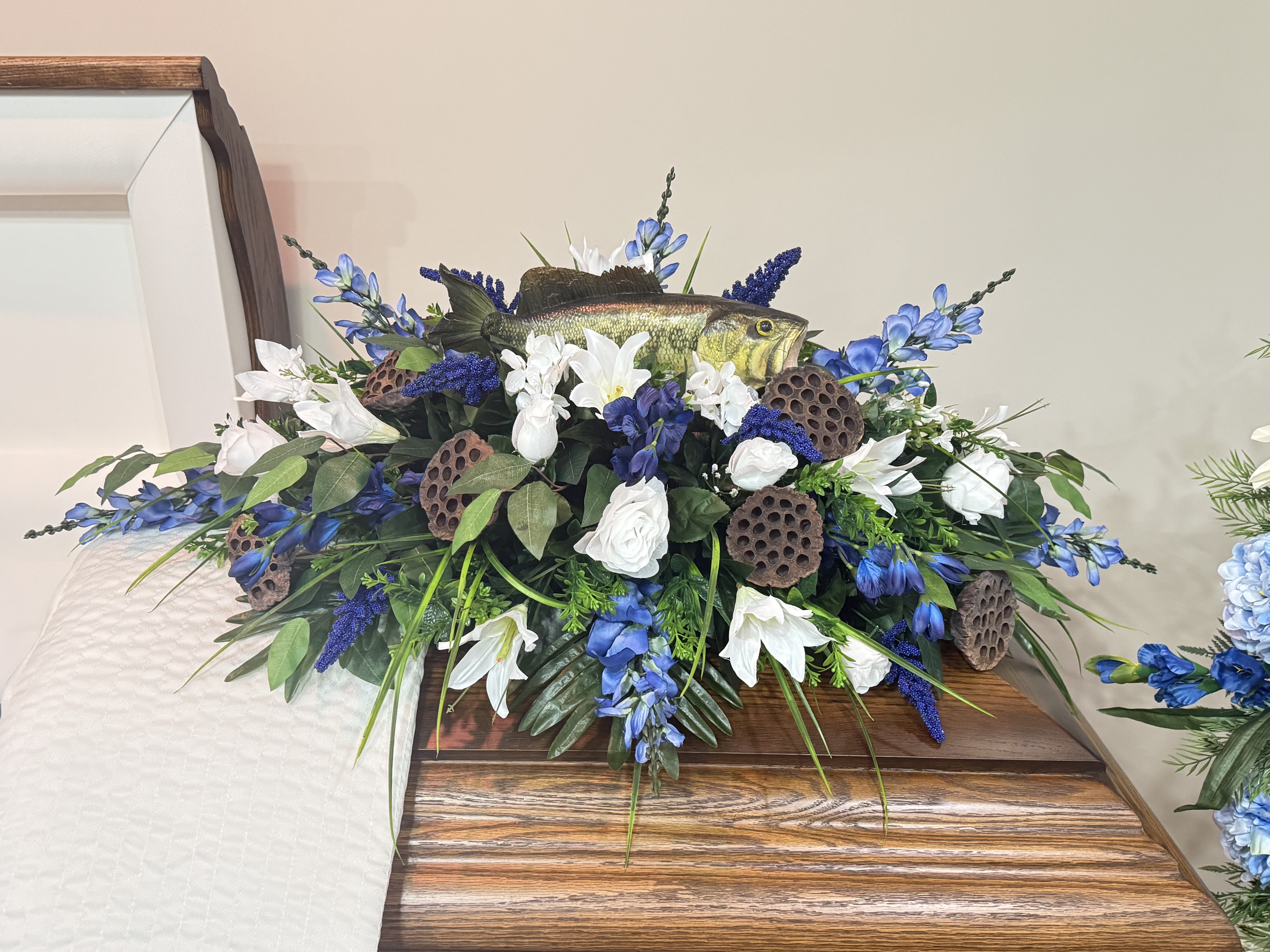 Hooked On Memories - Silk/Fresh Casket Spray  - Hooked on Memories is a special tribute for the one whose favorite days were spent on the water. Designed with white and blue blooms, natural lotus pods, and lush greenery, this casket spray features a lifelike largemouth bass at the center — a perfect symbol of their love for fishing and the outdoors.  Whether crafted in fresh flowers or lasting silk, this arrangement honors a life filled with peaceful moments, cast lines, and cherished memories that will never be forgotten.  Fresh or Silk Available Call For Custom Colors or Flower Requests (870) 352-0273