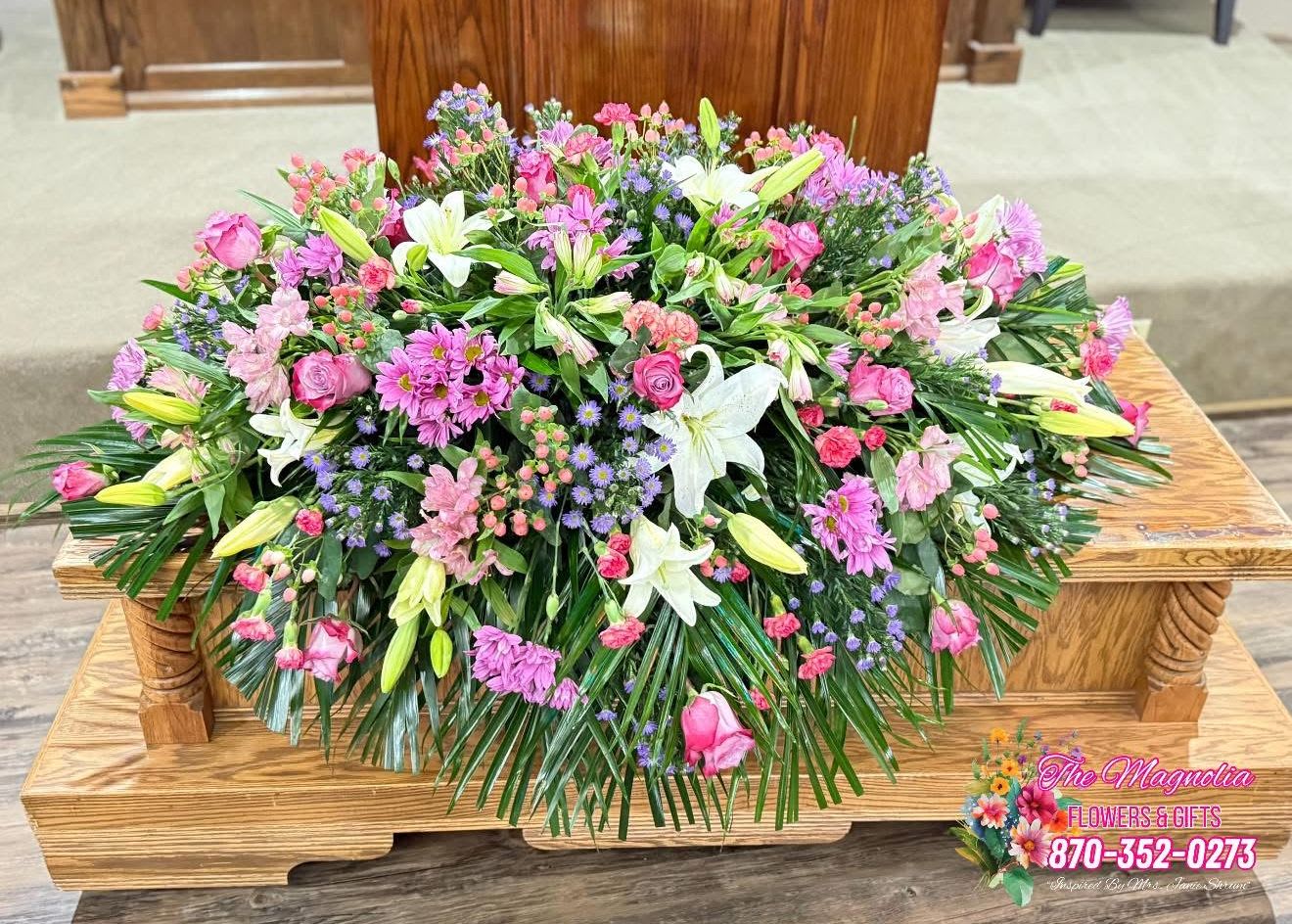 In Bloom Forever - Casket Spray - In Bloom Forever is a graceful and feminine casket spray, filled with soft shades of purple, white, and pink. Featuring elegant white lilies, purple roses, dainty daisies, pink alstroemeria, and lush cat palm greenery, this arrangement beautifully honors a life filled with love, kindness, and quiet strength.  Designed to feel like a blooming garden, this tribute is a reminder that the love they gave continues to grow in the hearts of those left behind.  Fresh or Silk Available Call For Custom Colors or Flower Requests (870) 352-0273