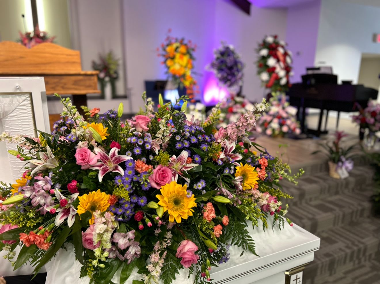 A Garden of Her Own - Casket Spray - A Garden of Her Own is a vibrant, garden-style tribute full of color, beauty, and life — just like she was. This arrangement features bold sunflowers, fragrant stargazer lilies, pink roses, larkspur, peach mini carnations, alstroemeria, solidago, and touches of purple montee, all nestled in flowing ivy and lush greenery.  Designed to feel like a hand-picked garden in full bloom, this spray celebrates her joyful spirit, her warmth, and the love she planted in the hearts of everyone she knew.  Fresh or Silk Available Call For Custom Colors or Flower Requests (870) 352-0273